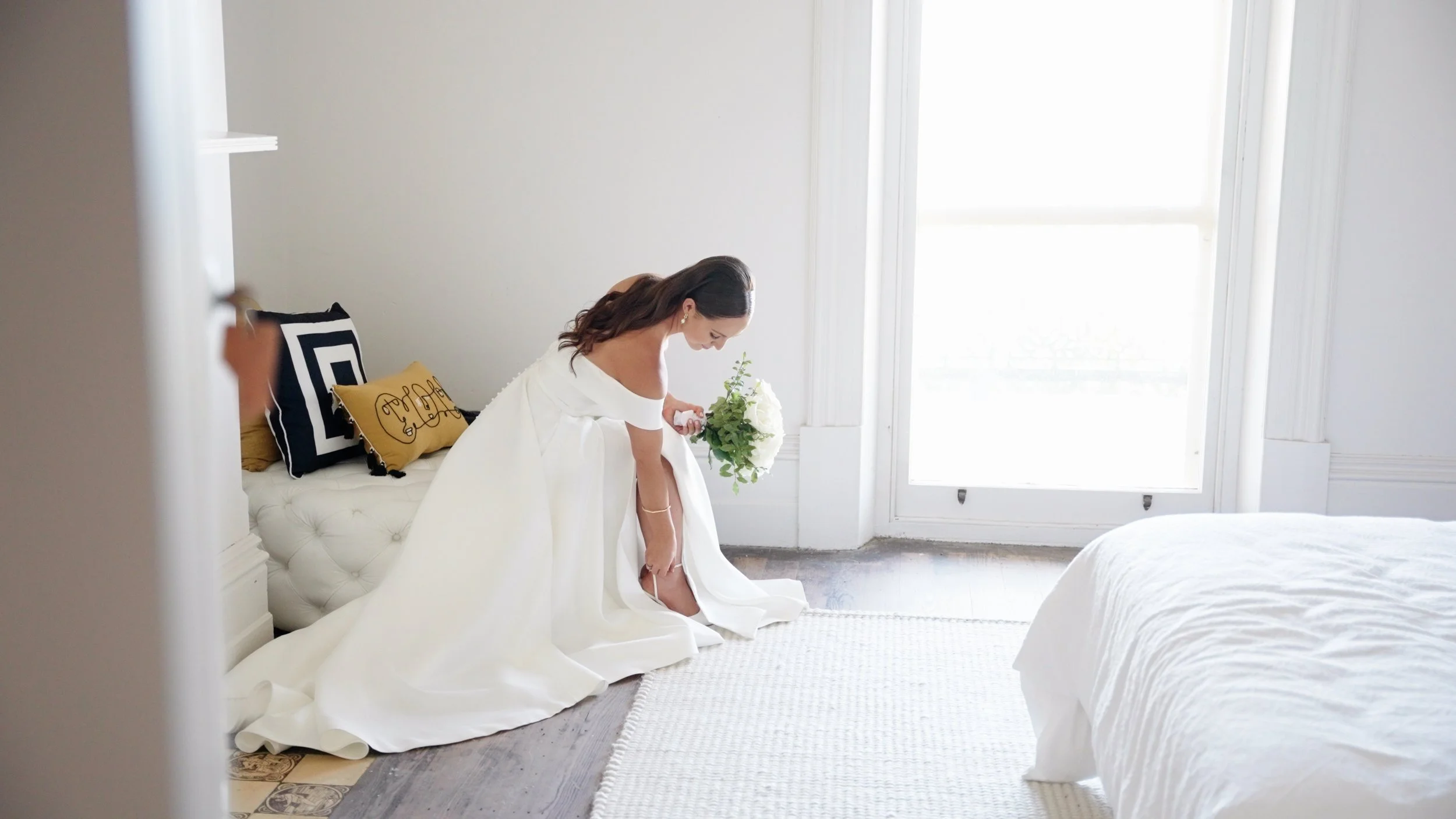 Bride in a white wedding dress bending down to put on her shoe in a bright room, holding a bouquet of white flowers.