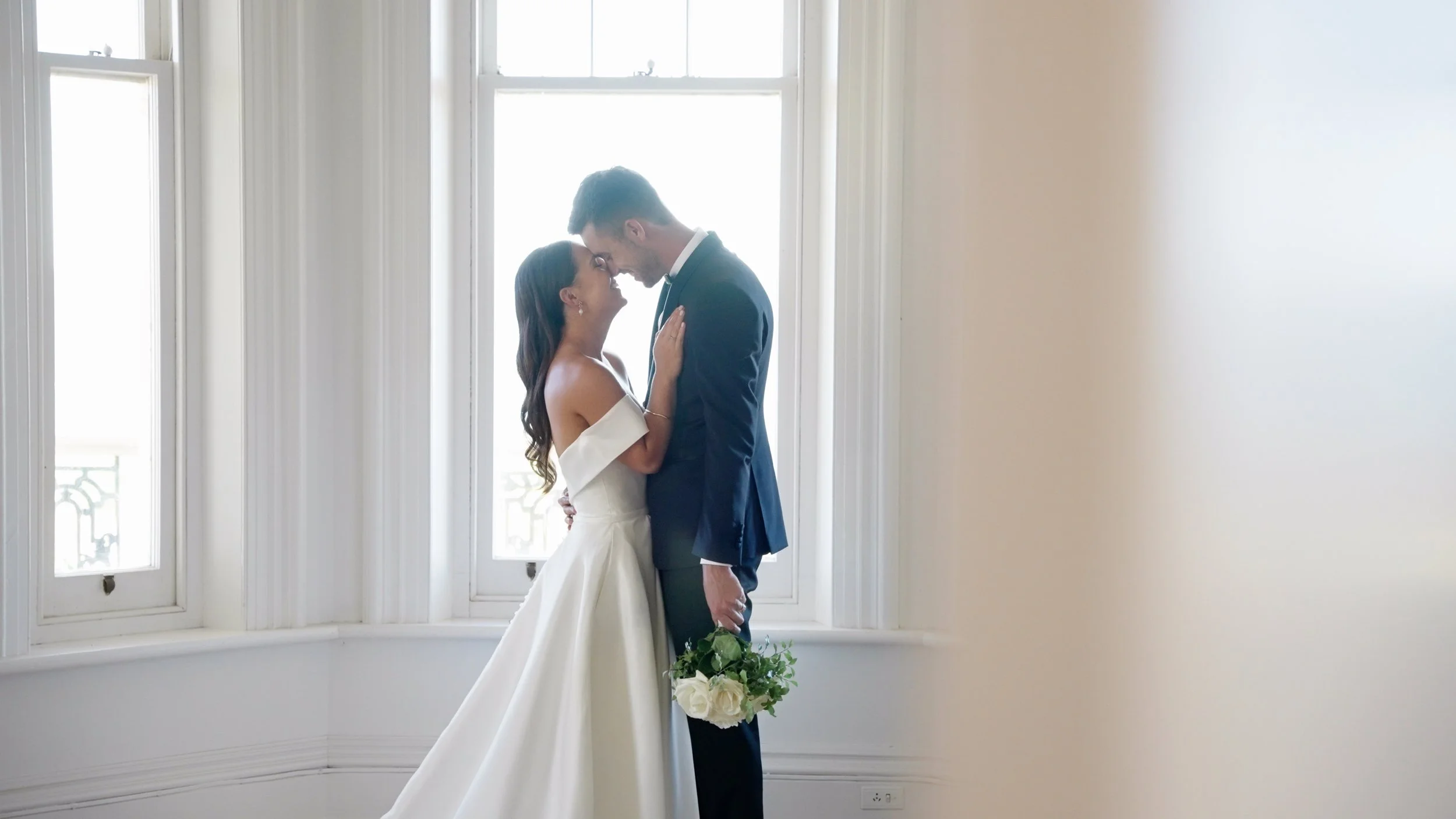 A bride and groom standing close together in front of a large window, holding each other and sharing a romantic moment, with the bride holding a bouquet of white roses.