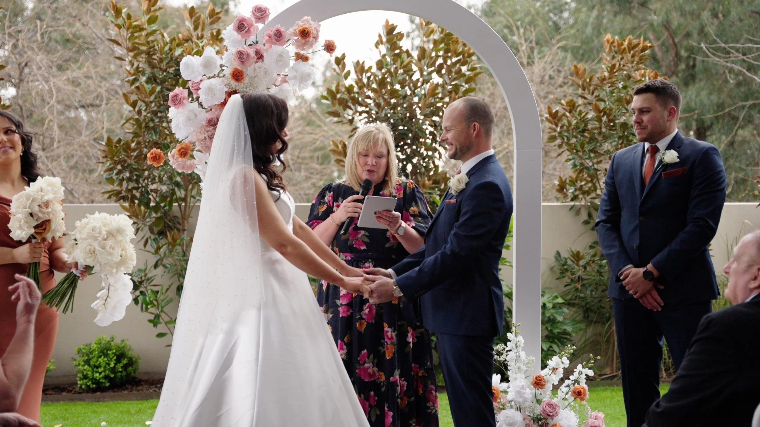 A bride and groom holding hands during their wedding ceremony outdoors, with an officiant speaking and witnesses holding bouquets, surrounded by floral arrangements and greenery.