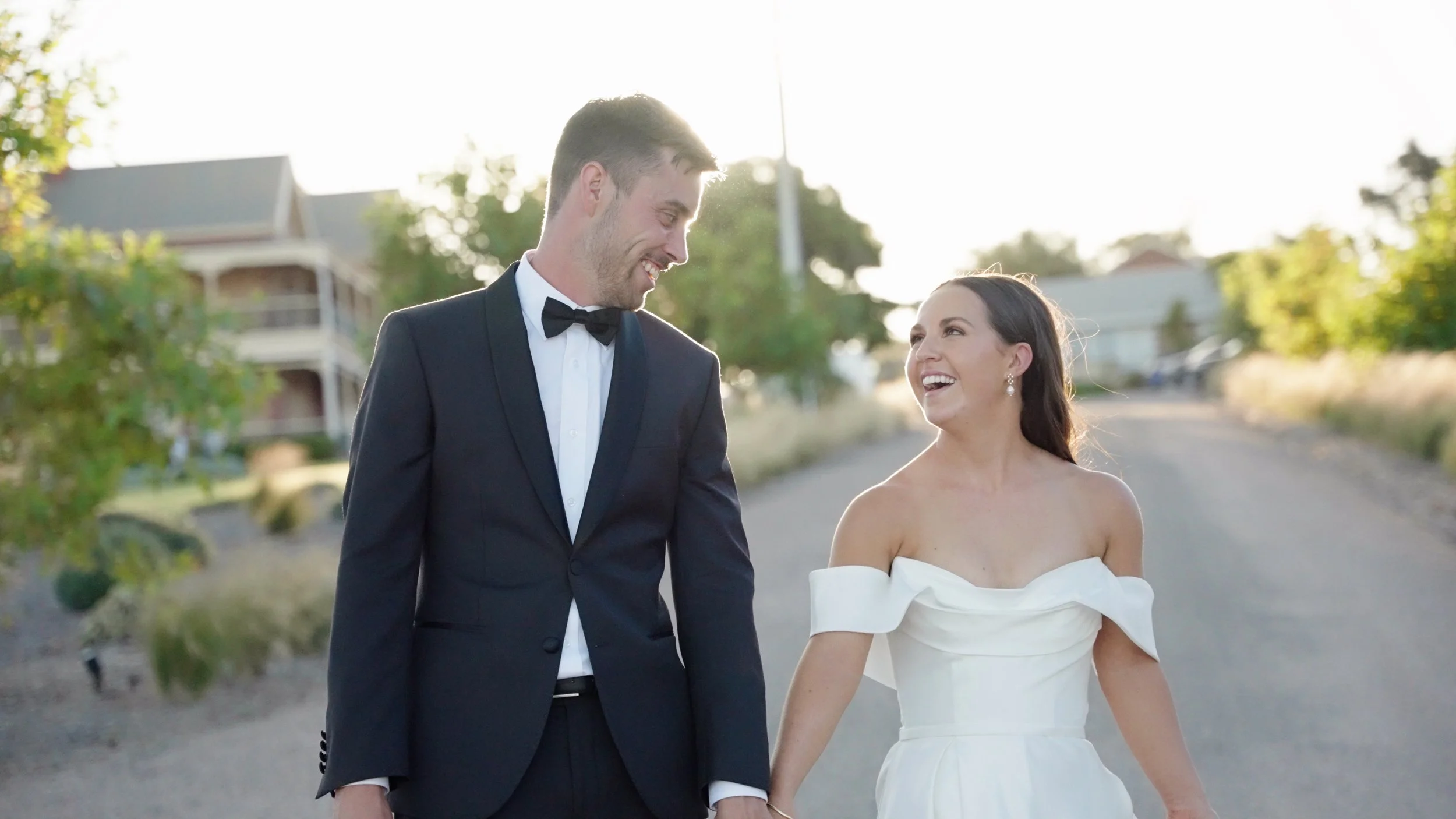 A bride and groom are walking hand in hand on a sunlit street, smiling and looking at each other. The groom is wearing a black tuxedo with a bow tie, and the bride is dressed in a white wedding gown with off-the-shoulder sleeves. They appear happy an