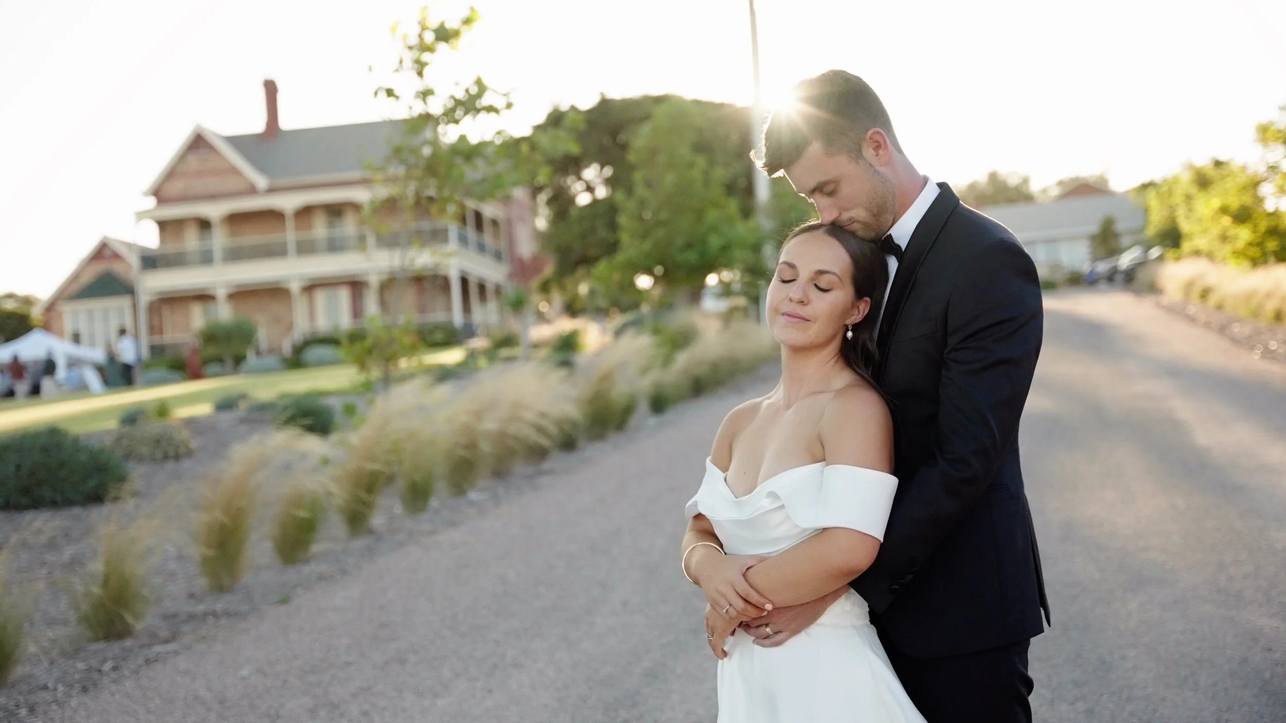 A couple dressed in wedding attire, embracing outdoors with sunlight shining behind them, and a house in the background.
