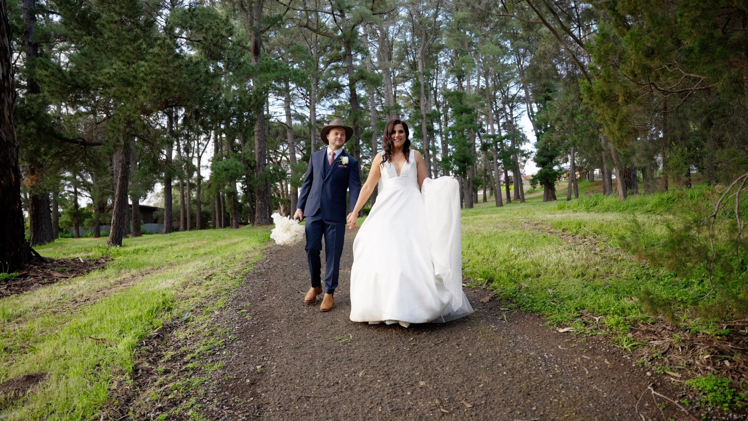 A newlywed couple walking hand in hand on a dirt path through a wooded area, smiling with trees surrounding them.