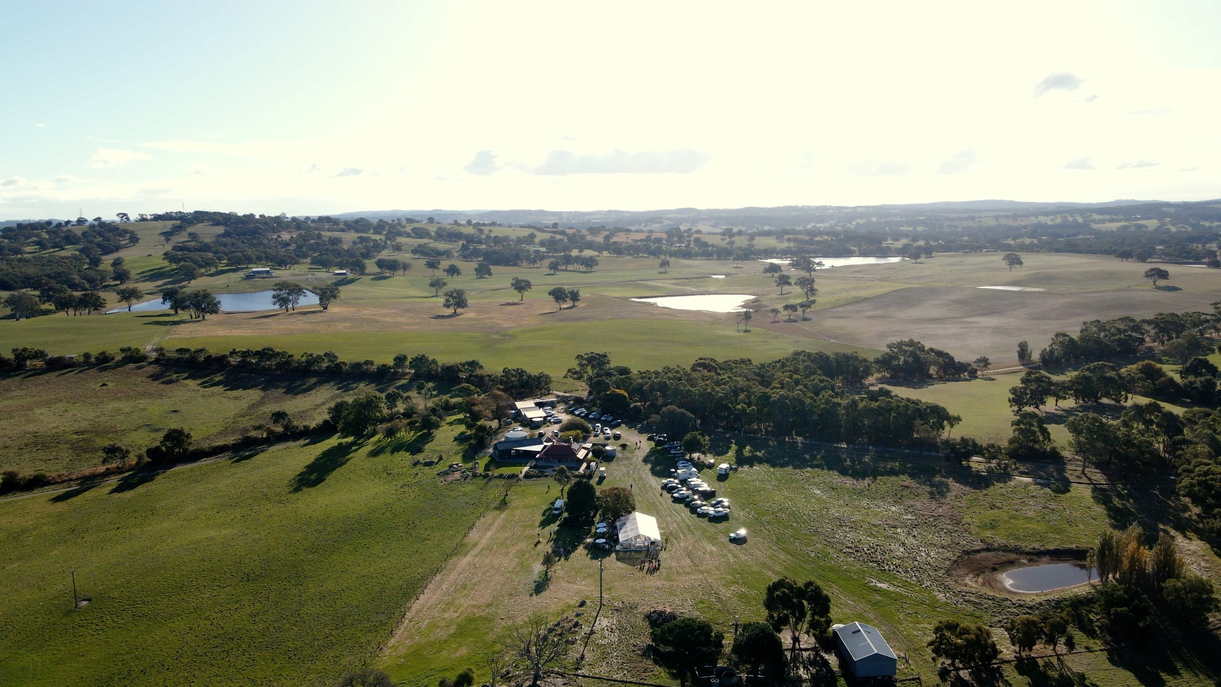 Aerial view of a rural landscape with fields, trees, water bodies, and a gathering area with cars and tents.