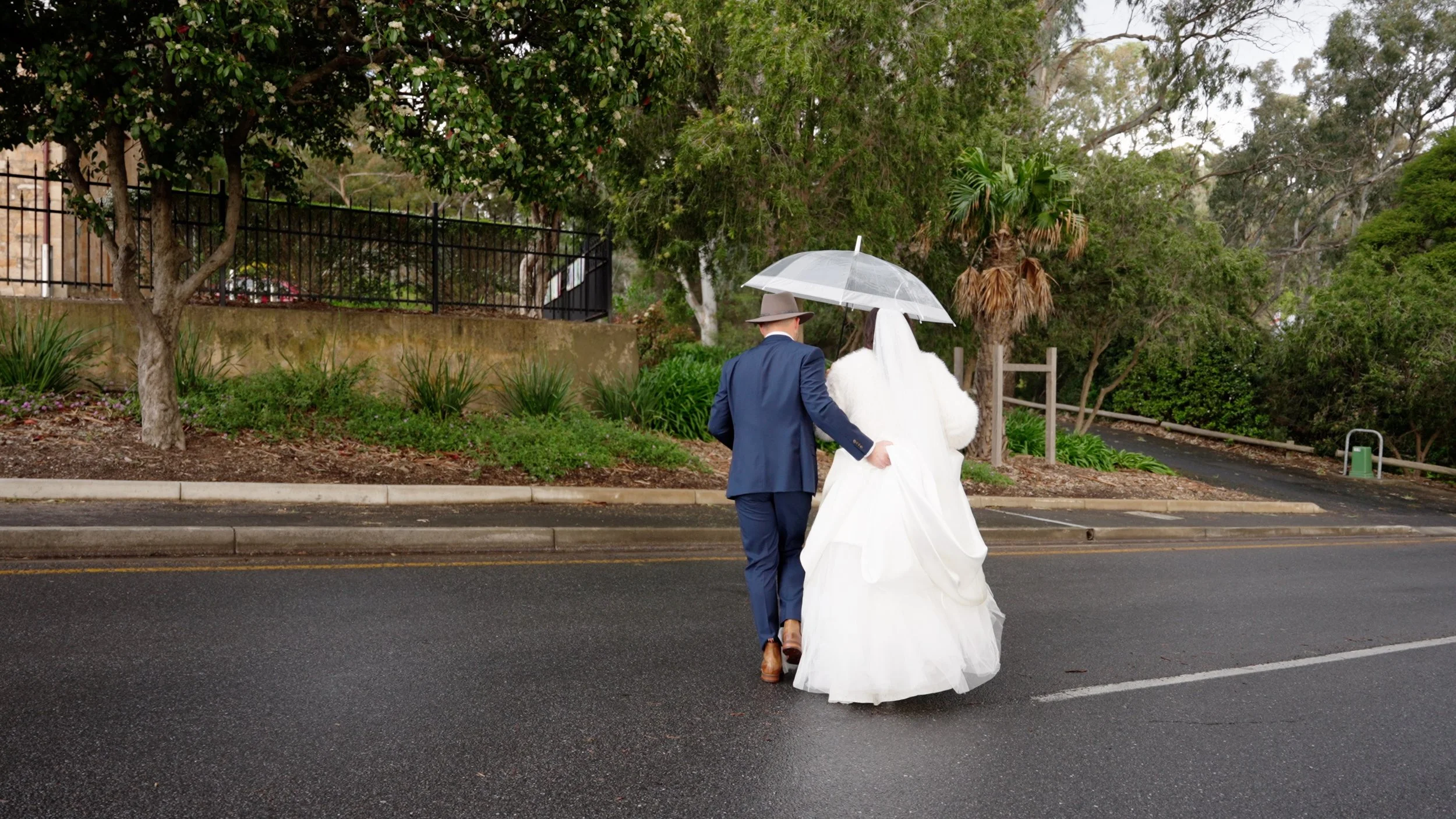 A bride and groom walking together in the rain, with the bride holding a transparent umbrella. The groom is dressed in a blue suit and brown shoes, and the bride is wearing a white wedding gown. The scene is set on a wet street with greenery and tree