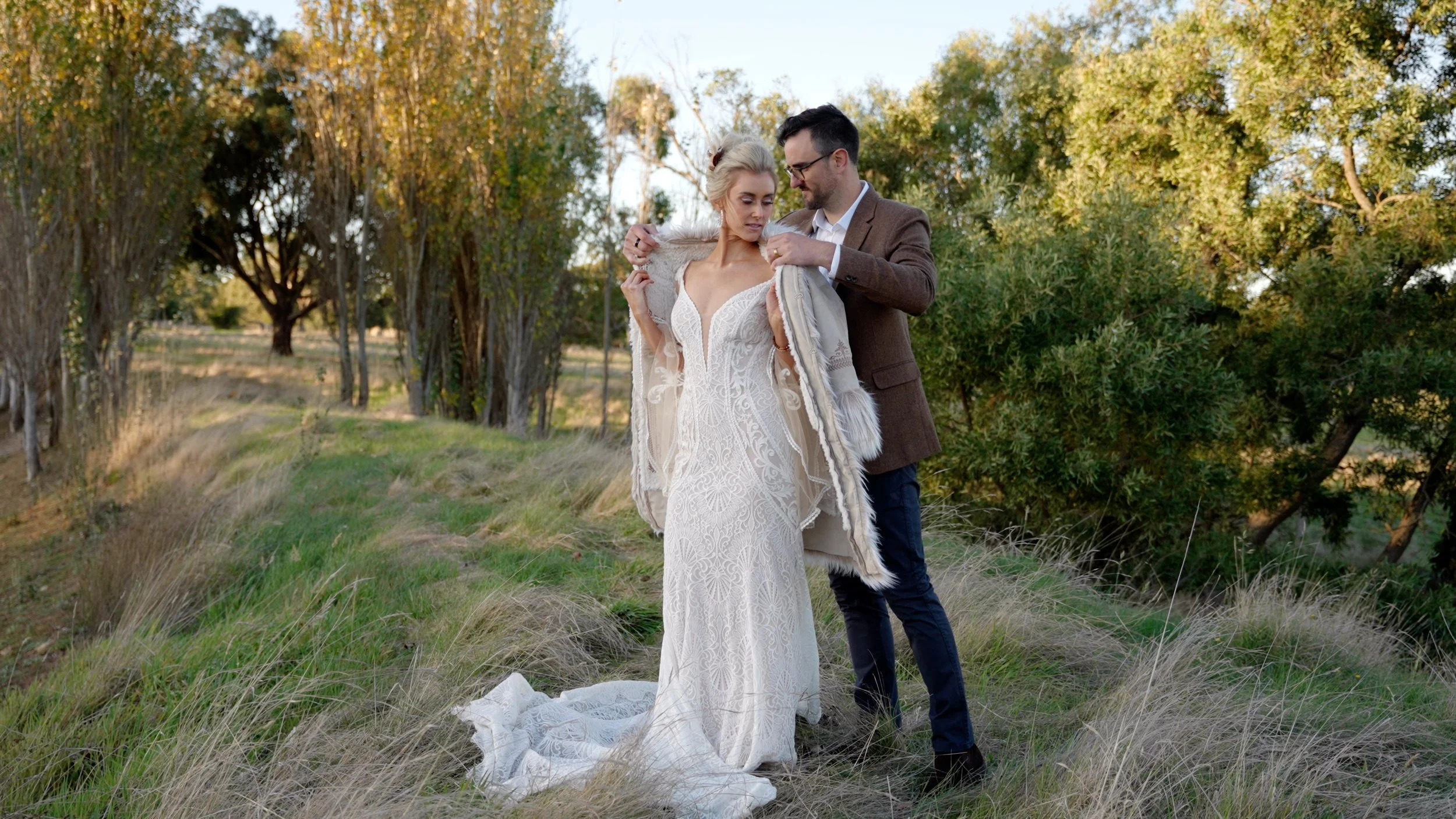 A couple stands outdoors in a grassy field, with trees in the background. The woman wears a white lace wedding dress and the man wears a brown suit jacket, as he adjusts her coat.