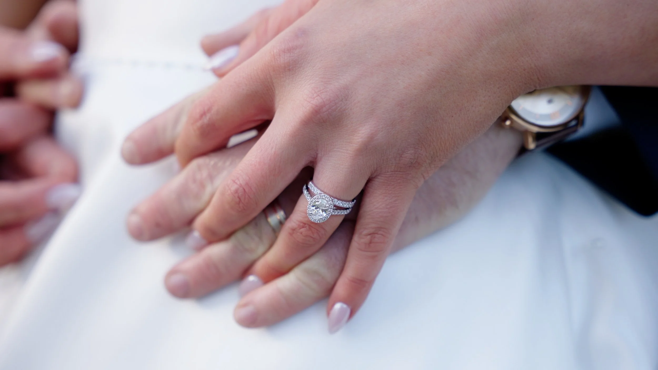 Close-up of a woman's hand resting on a man's hand, showing a diamond engagement ring and wedding band, with a wristwatch visible on the woman's wrist.