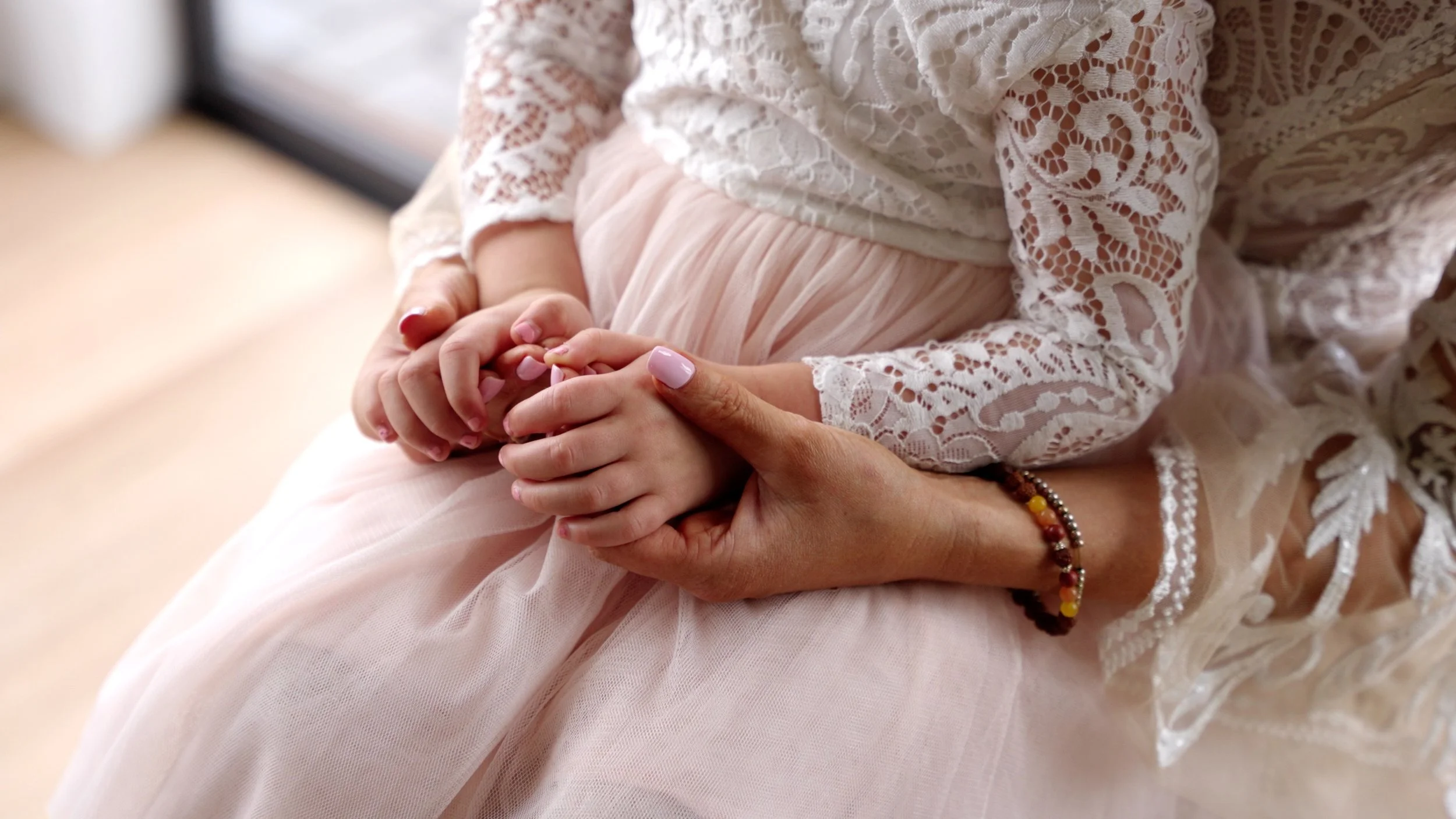 A woman holding a child's hands gently, with both wearing light-colored, lacy clothing.