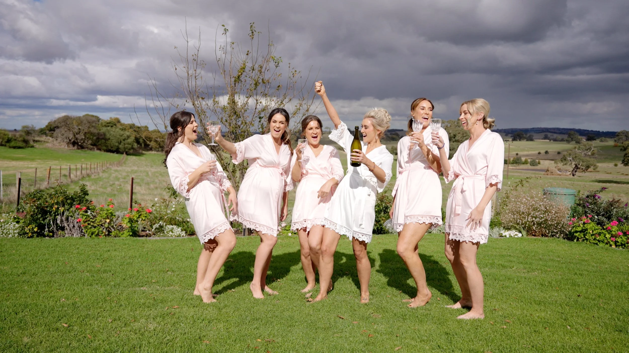 Seven women in matching white dresses celebrating outdoors on a grassy field with wine glasses, one holding a bottle, under a cloudy sky.