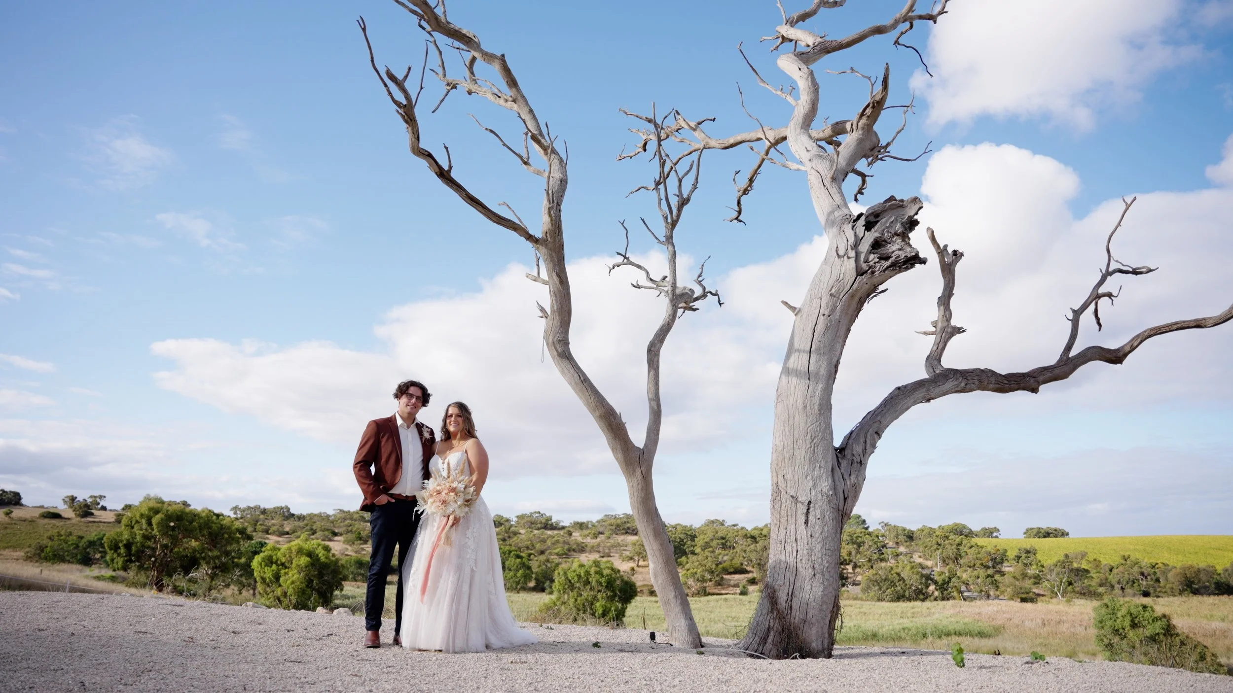 A bride and groom standing together outdoors in a rural landscape with a tall, leafless tree, green fields, and a partly cloudy sky in the background.