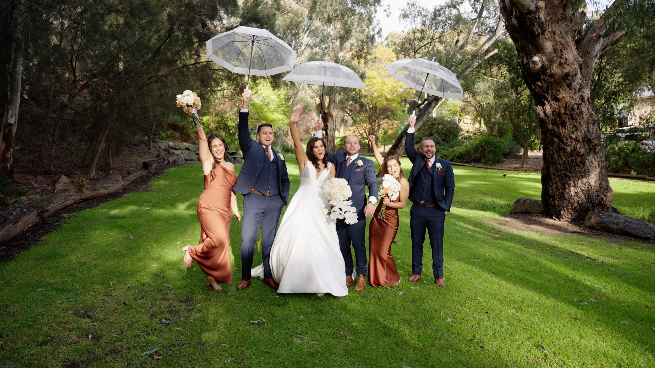 A wedding party celebrating outdoors on a grassy area with trees, holding umbrellas and raising their hands in joy, including a bride in a white dress, and bridesmaids and groomsmen in formal attire.