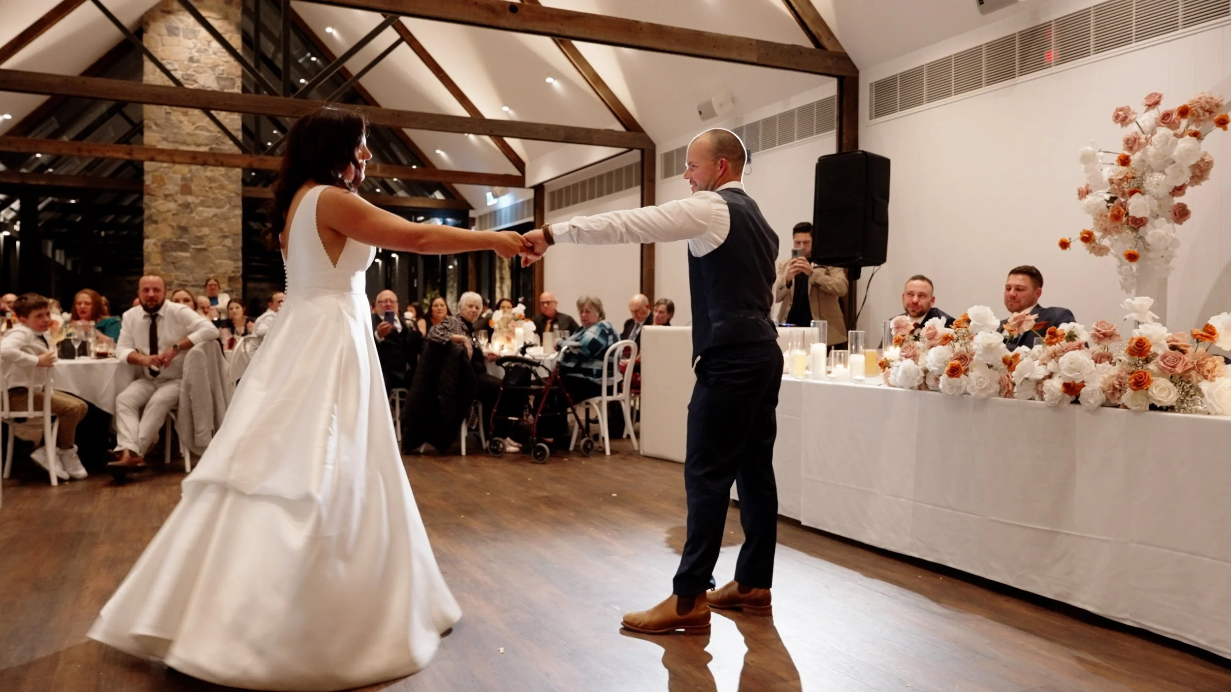 A bride and groom are dancing together at their wedding reception, holding hands and smiling, with seated guests watching in the background and floral decorations on the table nearby.