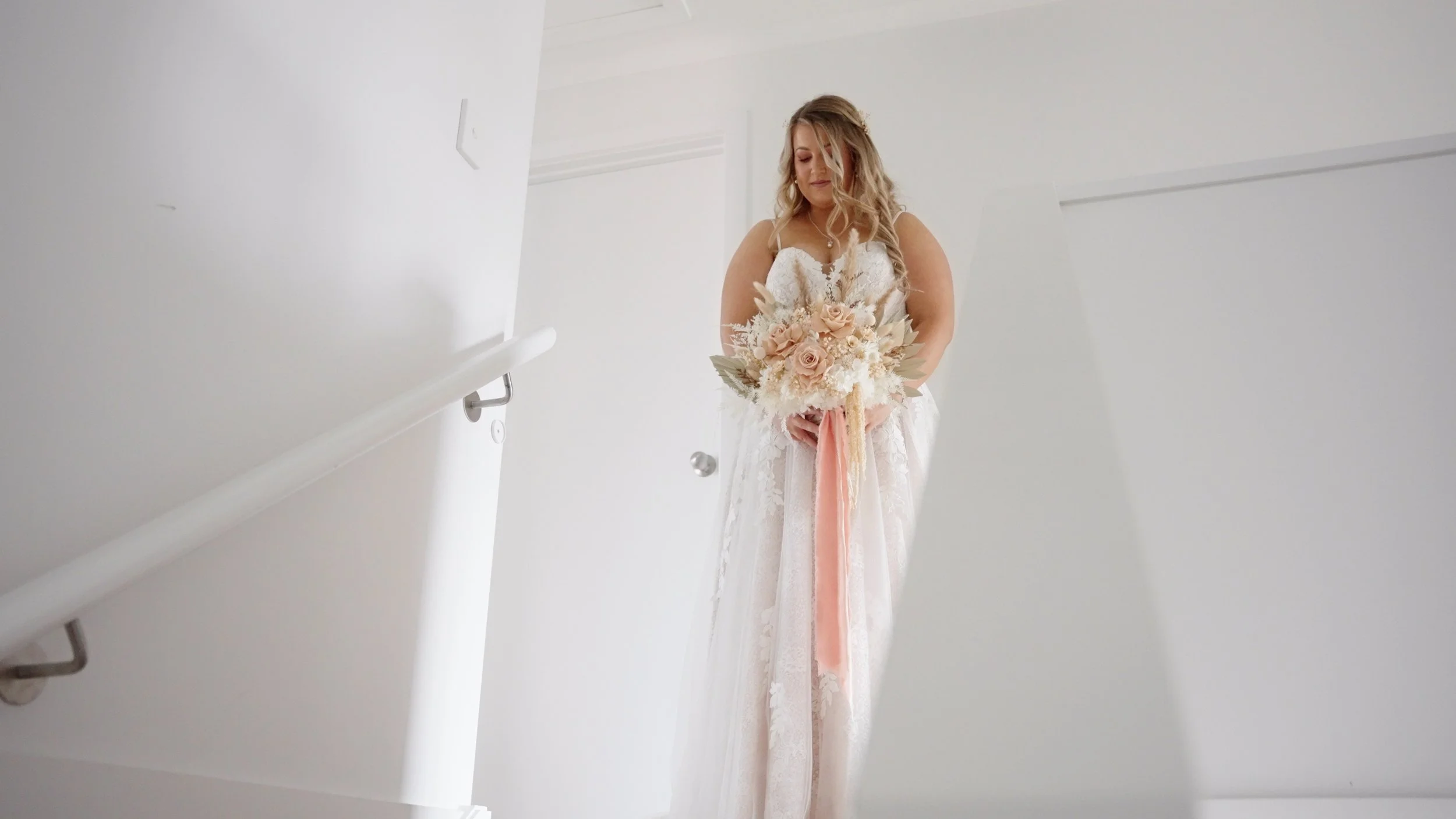 Bride in white lace dress holding a bouquet of roses and dried flowers, standing on a staircase with white walls.