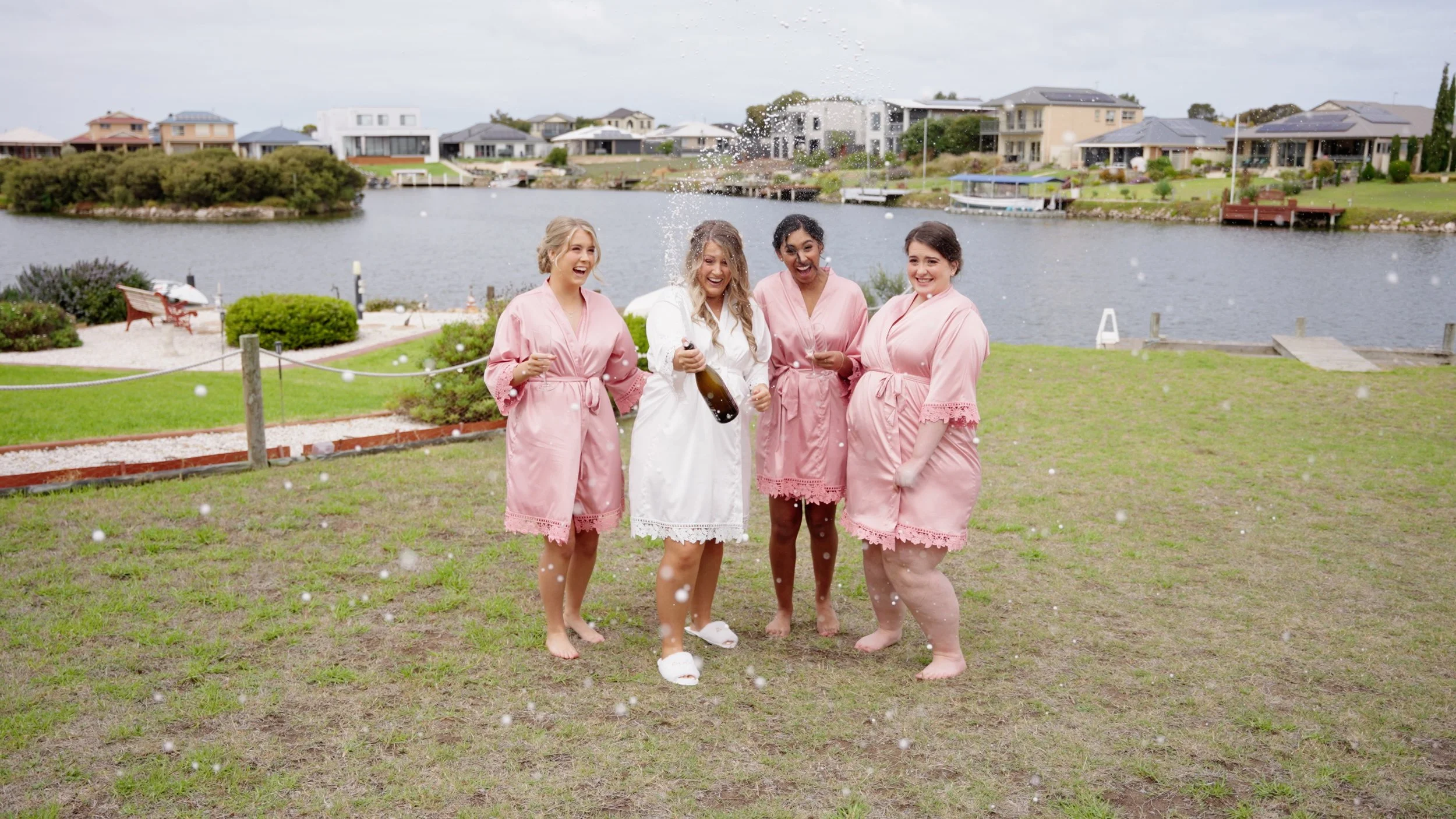Four women in pink and white robes celebrating by a lake, spraying champagne, with houses in the background.