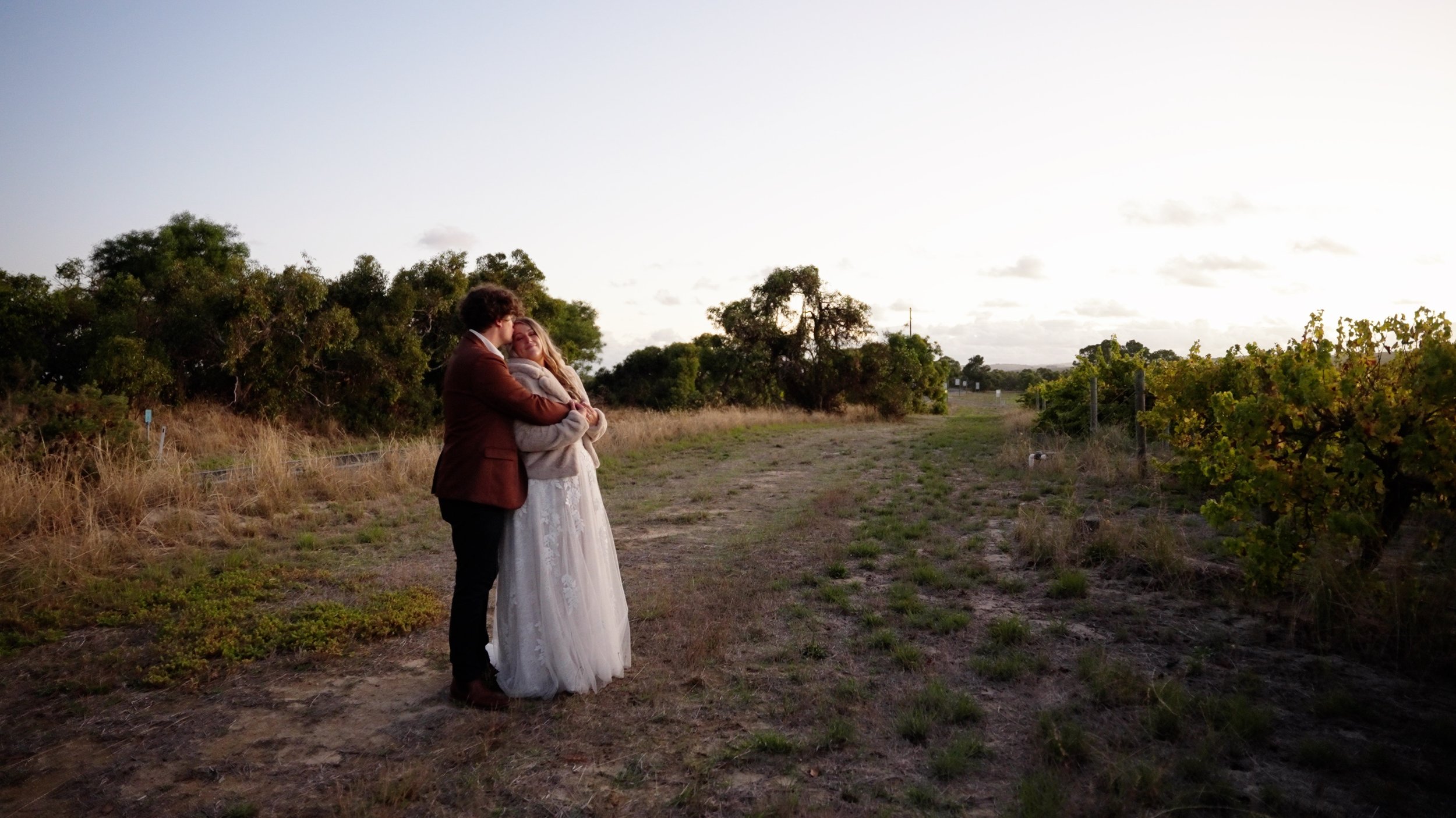 A couple embracing in a field at sunset, with trees and a dirt path in the background.