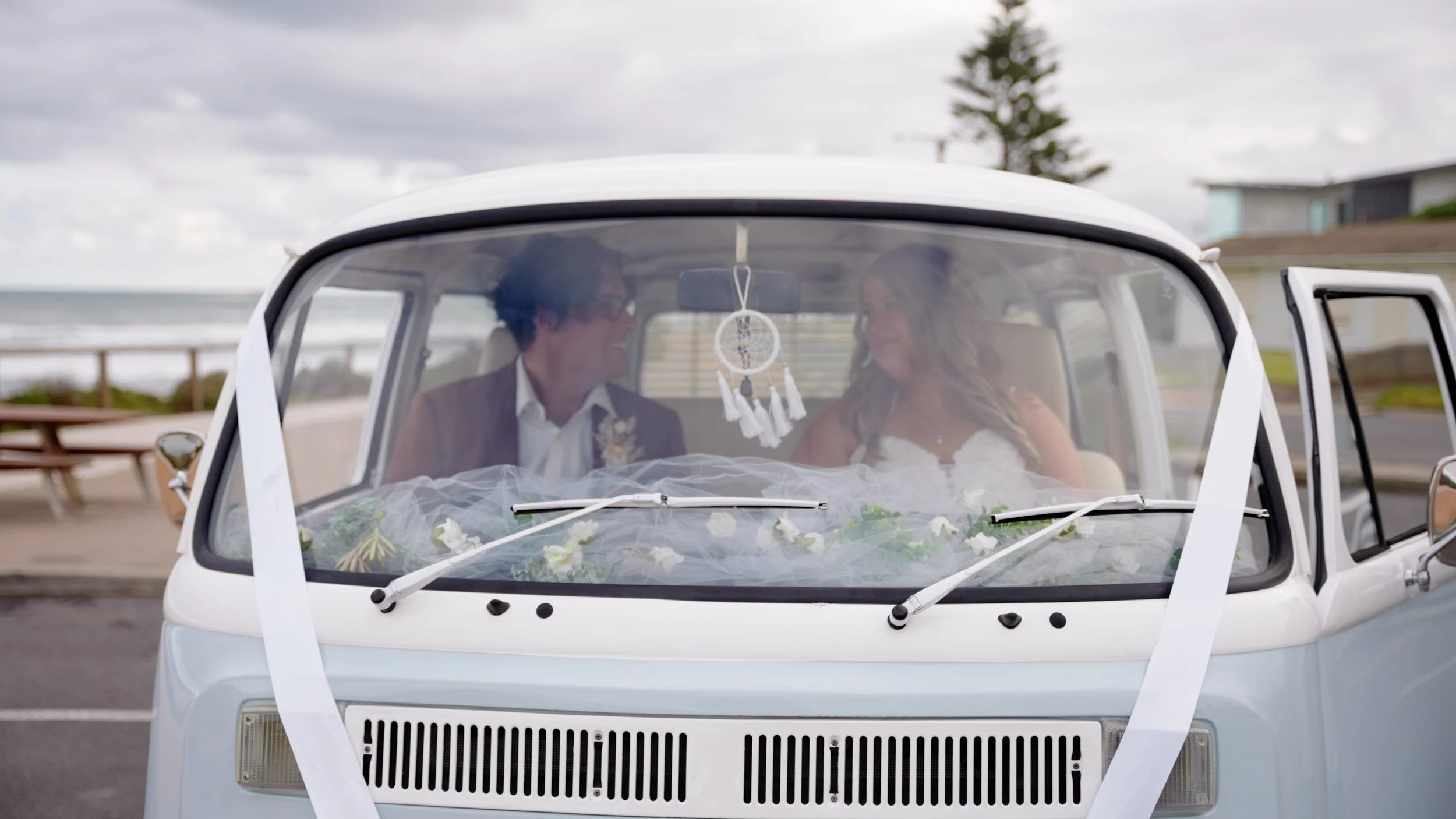 Couple in wedding attire sitting inside a decorated vintage van at an outdoor location near the beach, smiling and looking at each other.