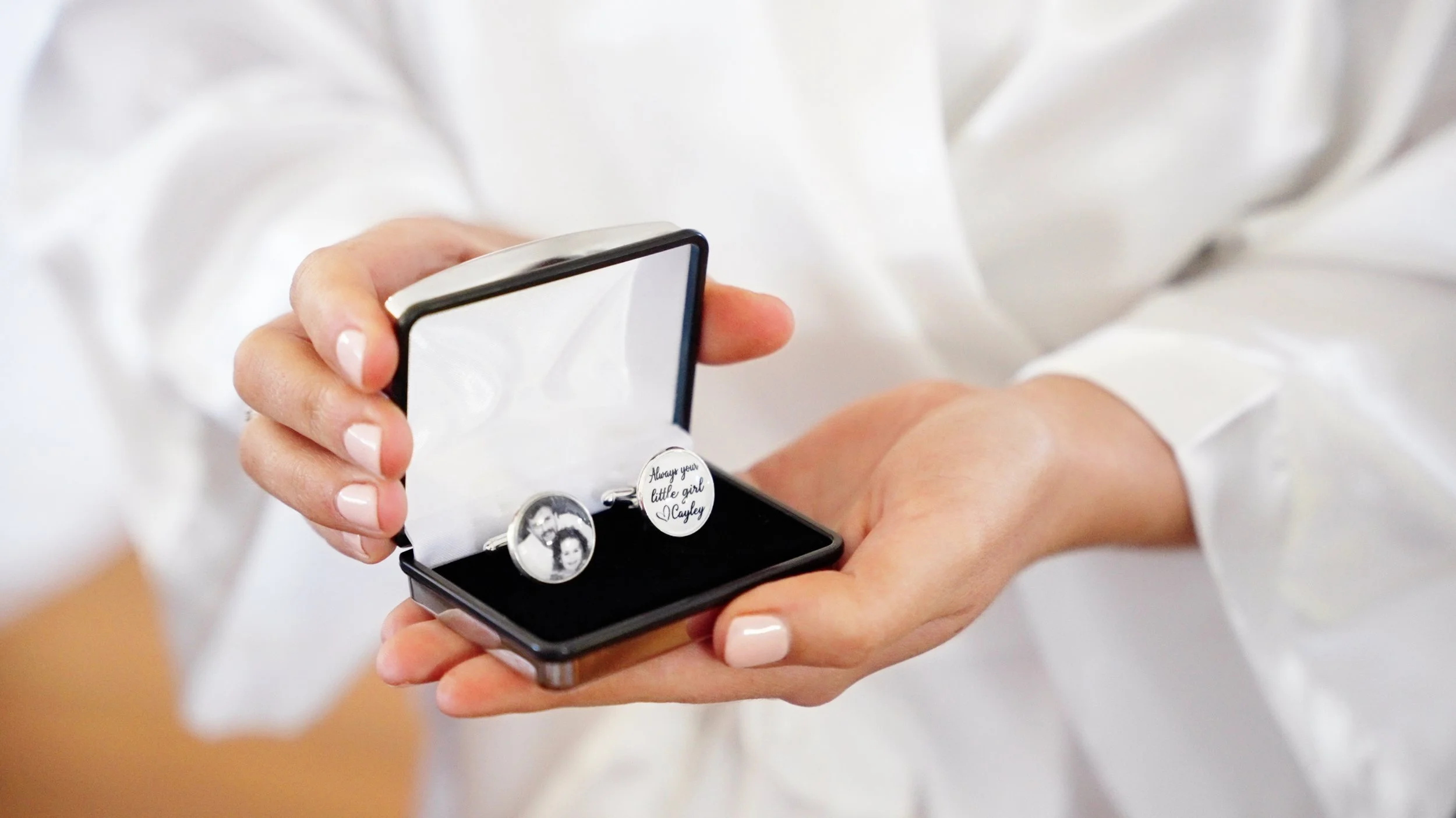 A person holding an open black jewelry box with cufflinks, one with a black and white photo and the other with a personalized message, inside.