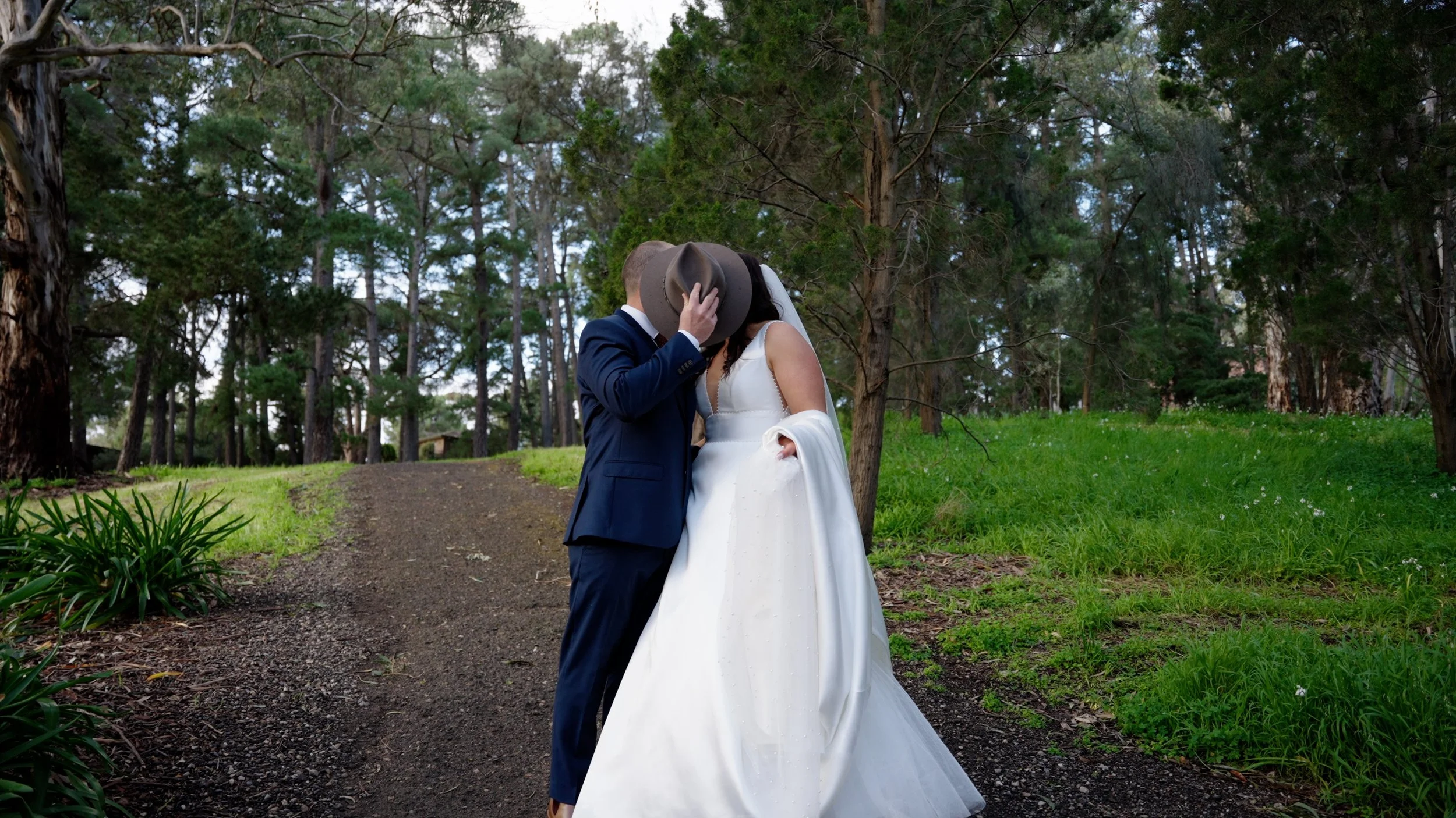 A bride and groom sharing a kiss outdoors in a wooded area, dressed in wedding attire, with the groom in a navy suit and the bride in a white wedding gown, holding her dress with one hand and tipping his hat with the other.