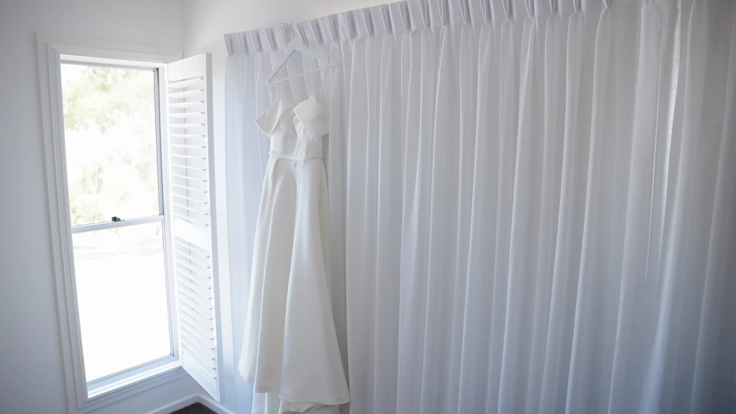 White wedding dress hanging on a white hanger near a window with shutters in a bright room.
