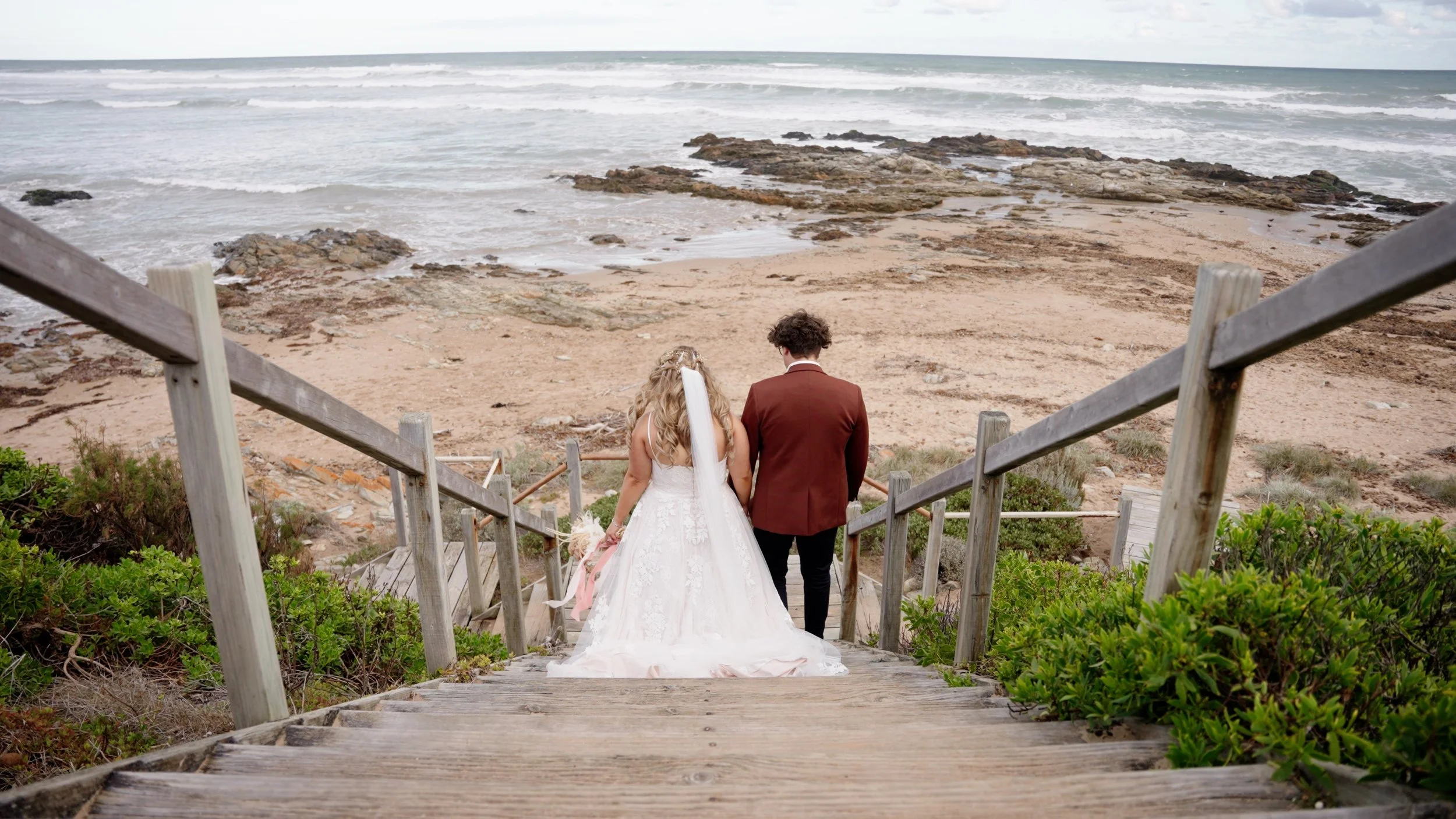 A bride and groom walk down a wooden staircase toward the beach, ocean waves, rocky shoreline, and cloudy sky in the background.