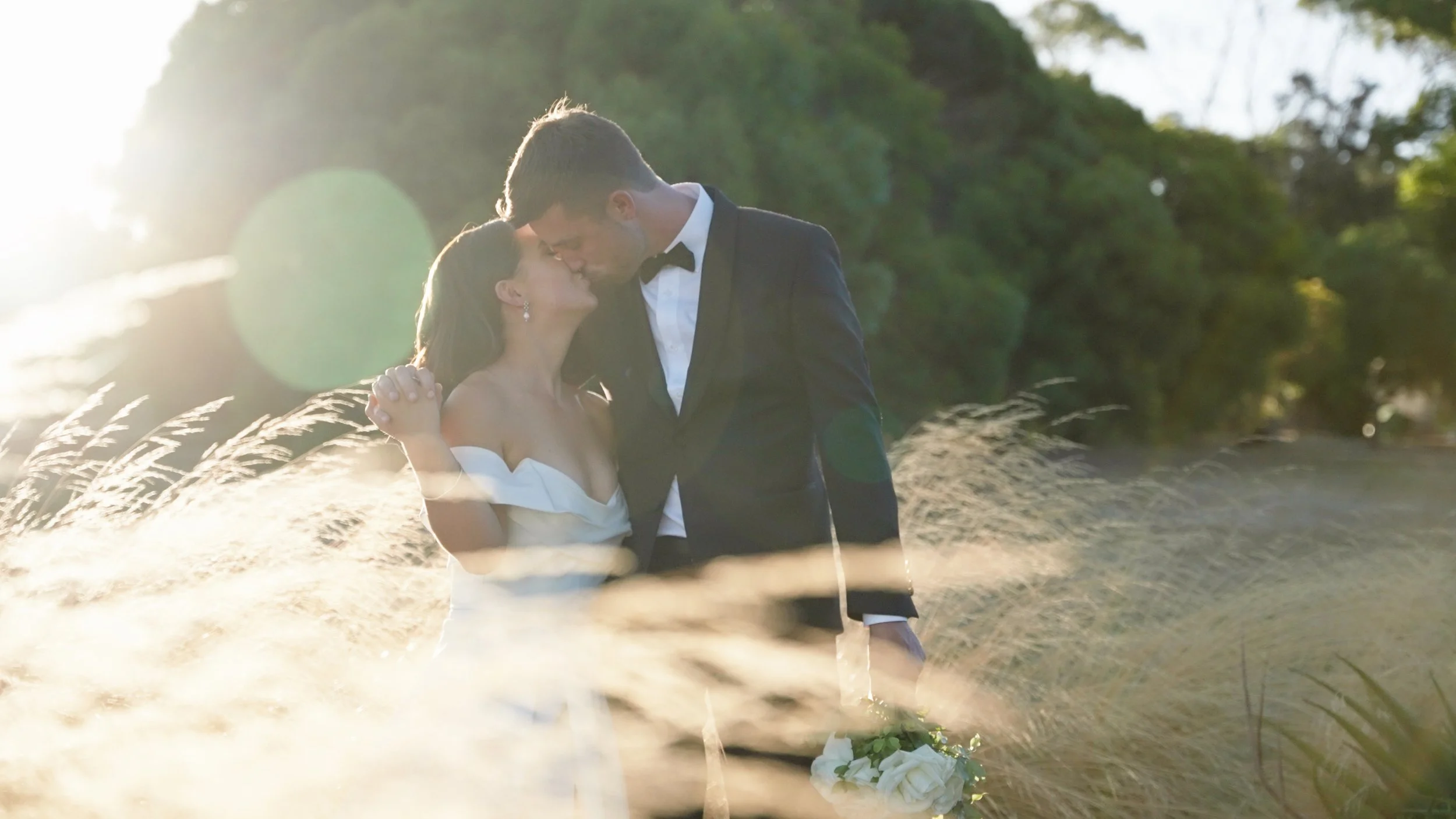 A couple in wedding attire sharing a kiss outdoors during sunset, with a natural landscape and grasses in the background.