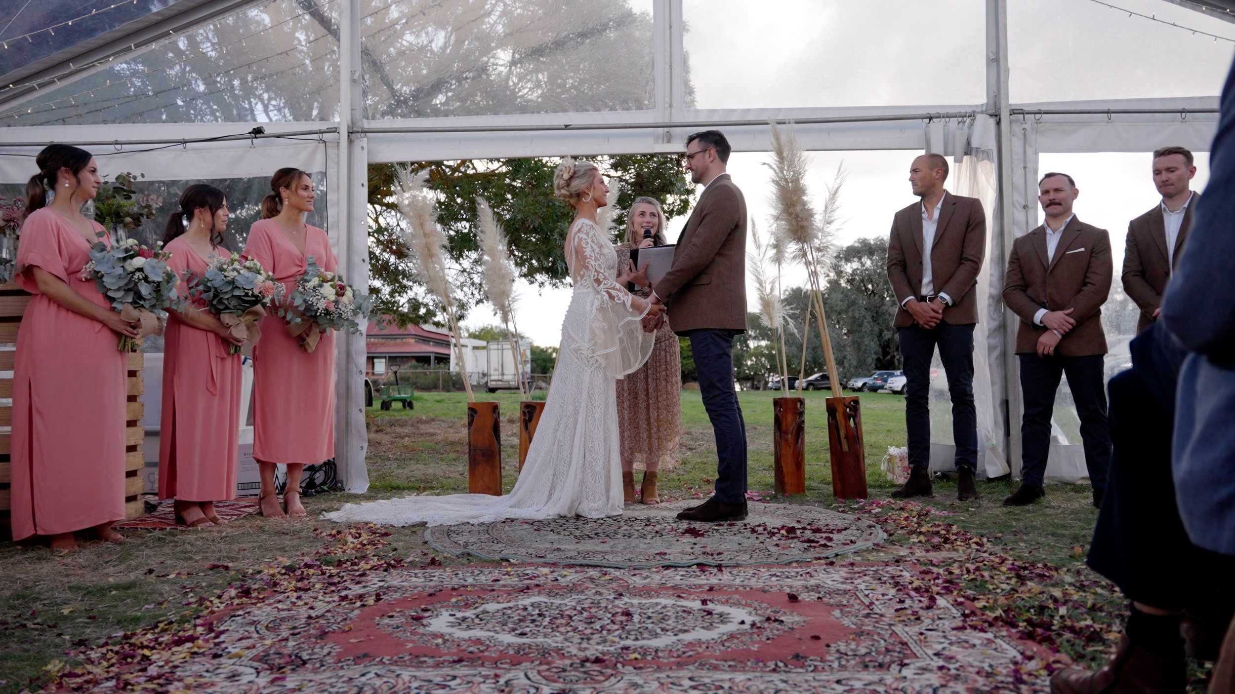 A wedding ceremony taking place outdoors inside a transparent tent, with the bride and groom holding hands and exchanging vows. The bride is wearing a white lace wedding dress, the groom a brown jacket, and they are facing each other. Four bridesmaid