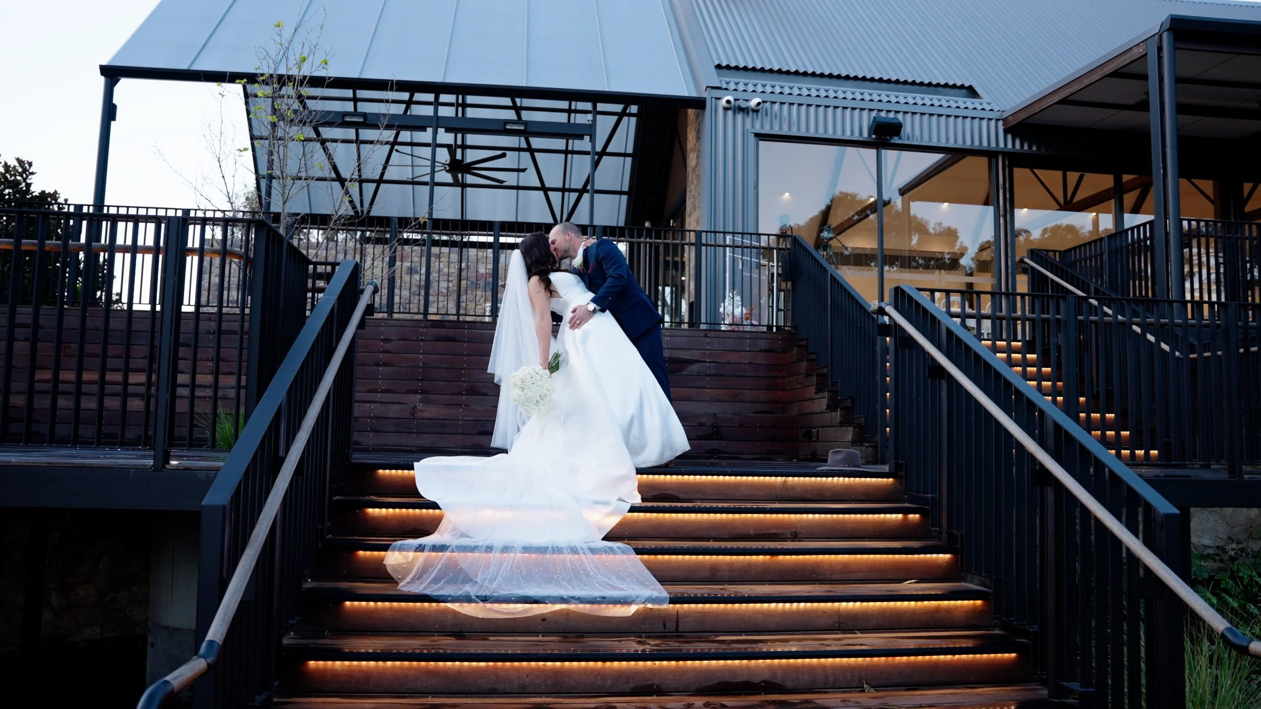 A bride and groom on wedding stairs, embracing and sharing a kiss, with a modern building in the background.
