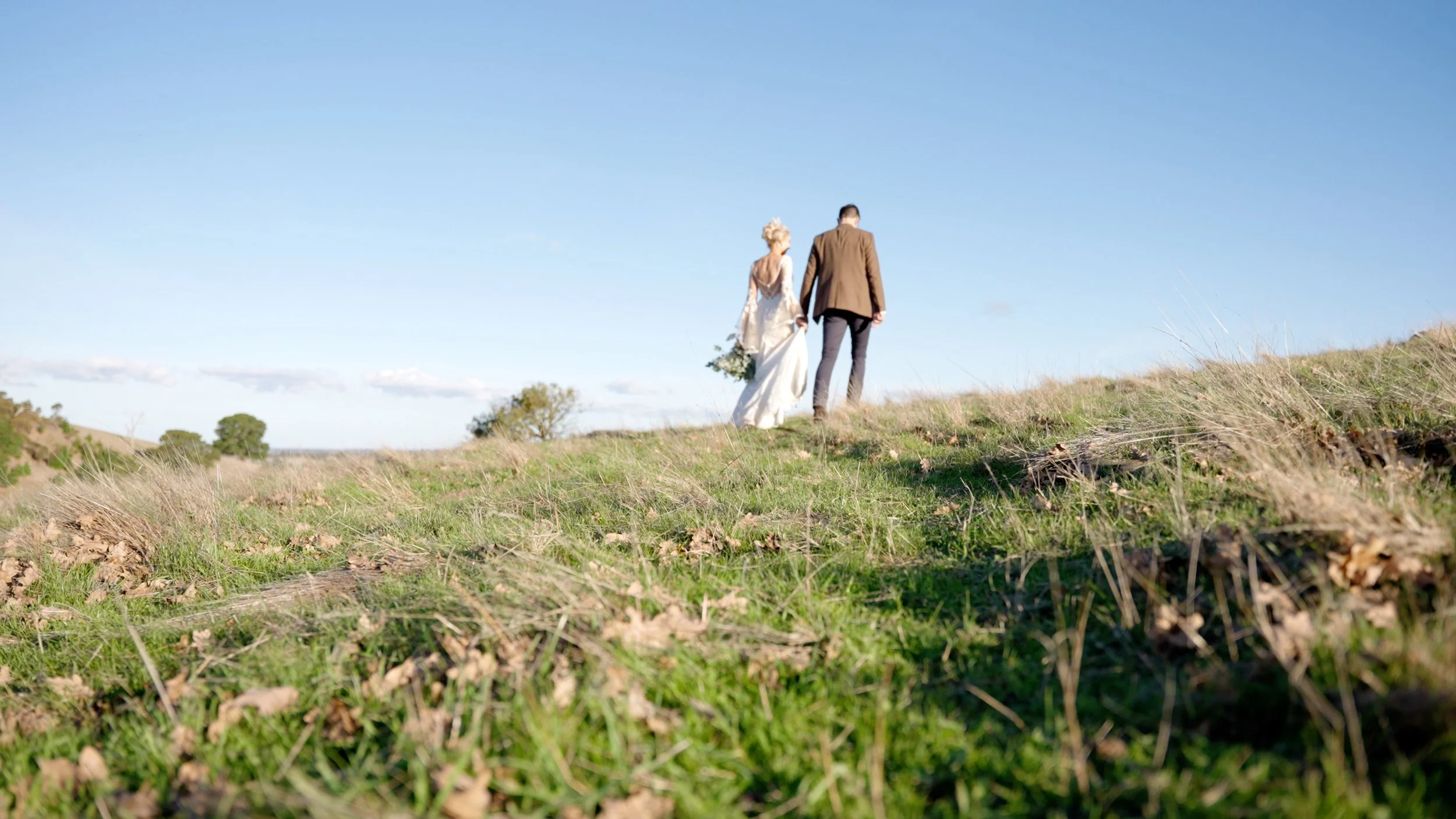 A bride and groom walking hand in hand on a grassy hill under a clear blue sky