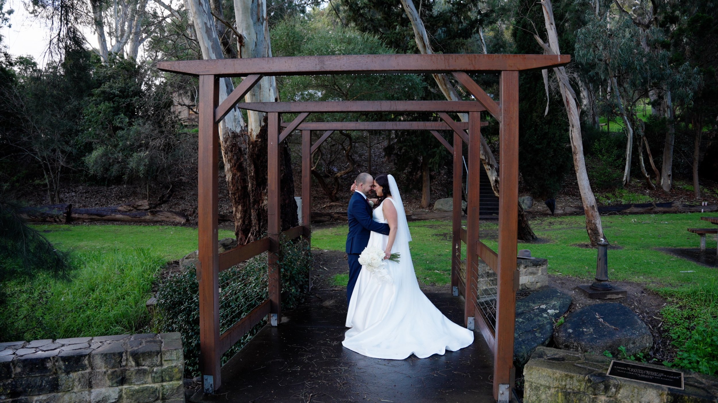 A bride and groom stand under a wooden arch, sharing an intimate moment in a park-like setting with trees and greenery.