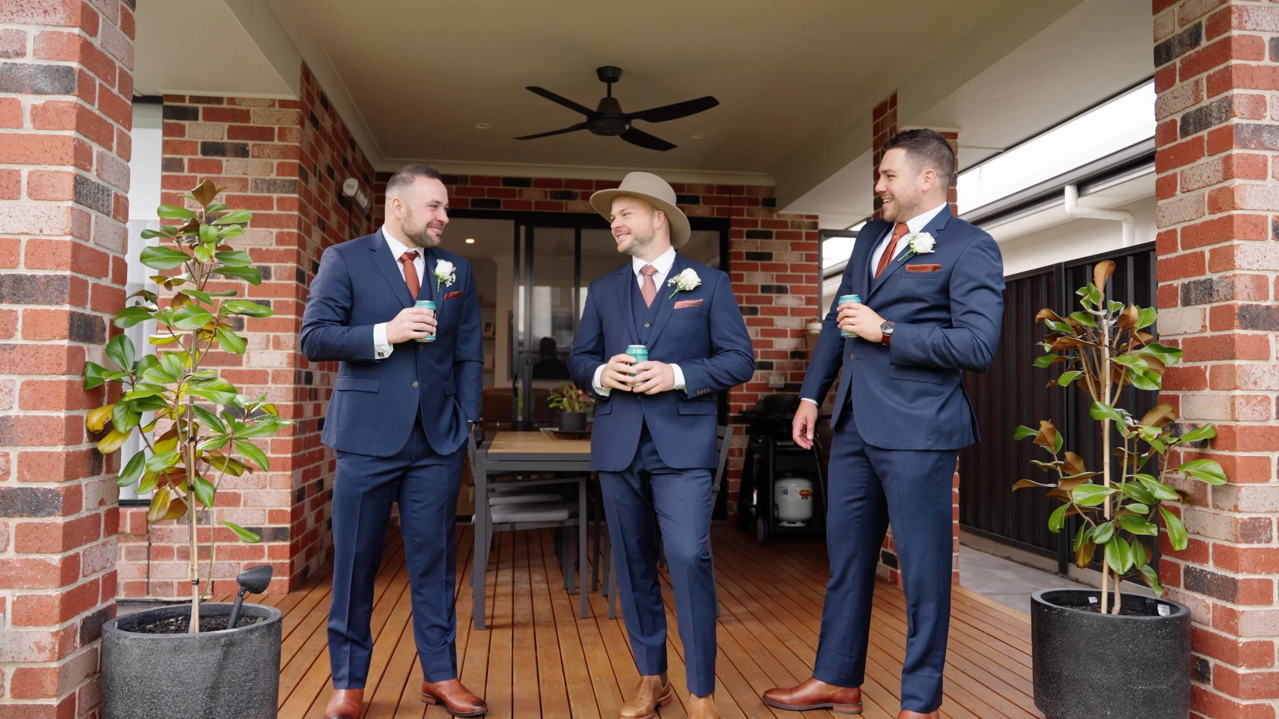 Three men dressed in blue suits, white shirts, and brown shoes standing on a wooden patio, holding drinks, and smiling at each other. The background features a brick wall, potted plants, and a ceiling fan.
