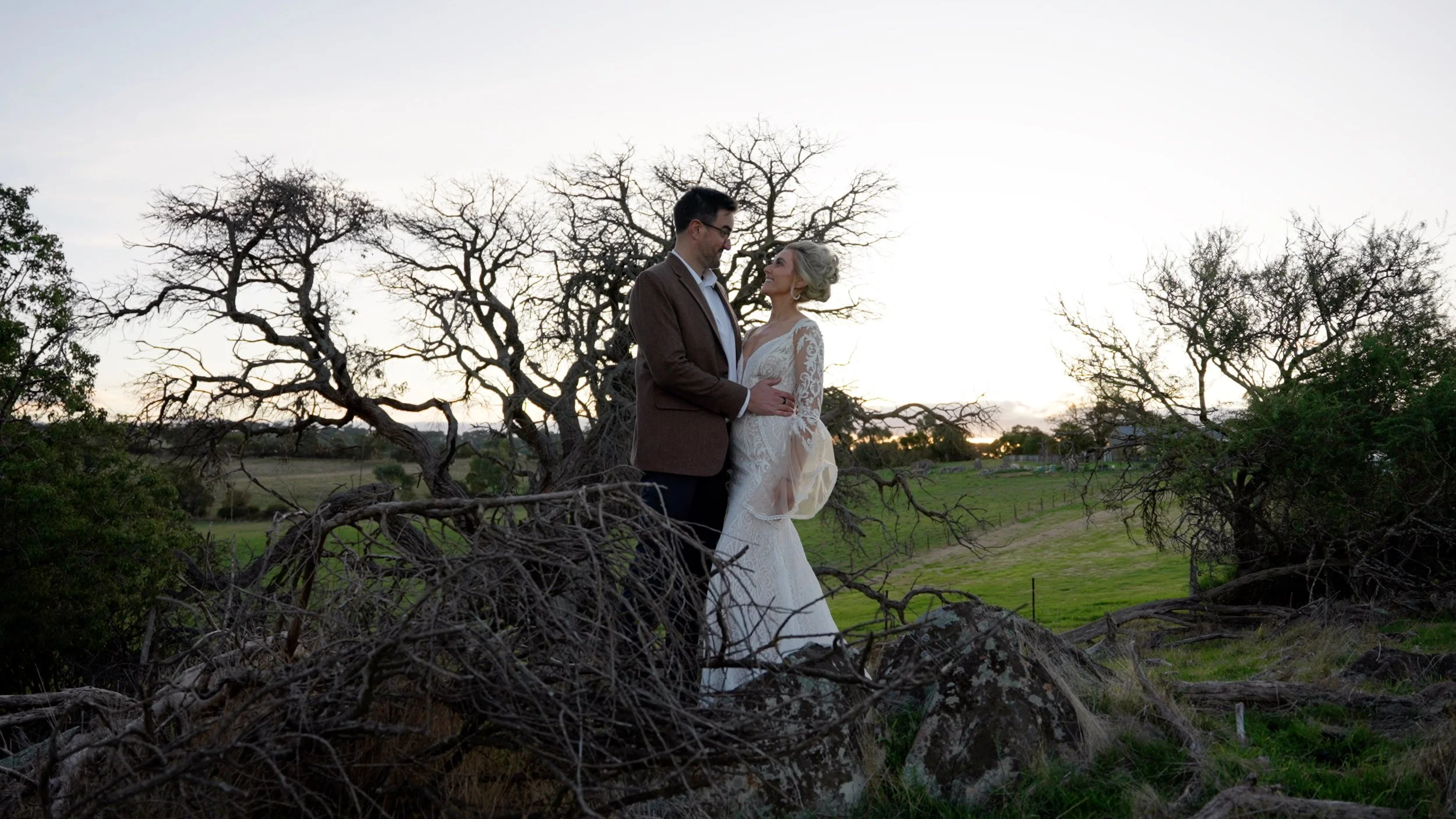 A bride and groom standing close together outdoors during sunset in a rural area with leafless trees and green grass.