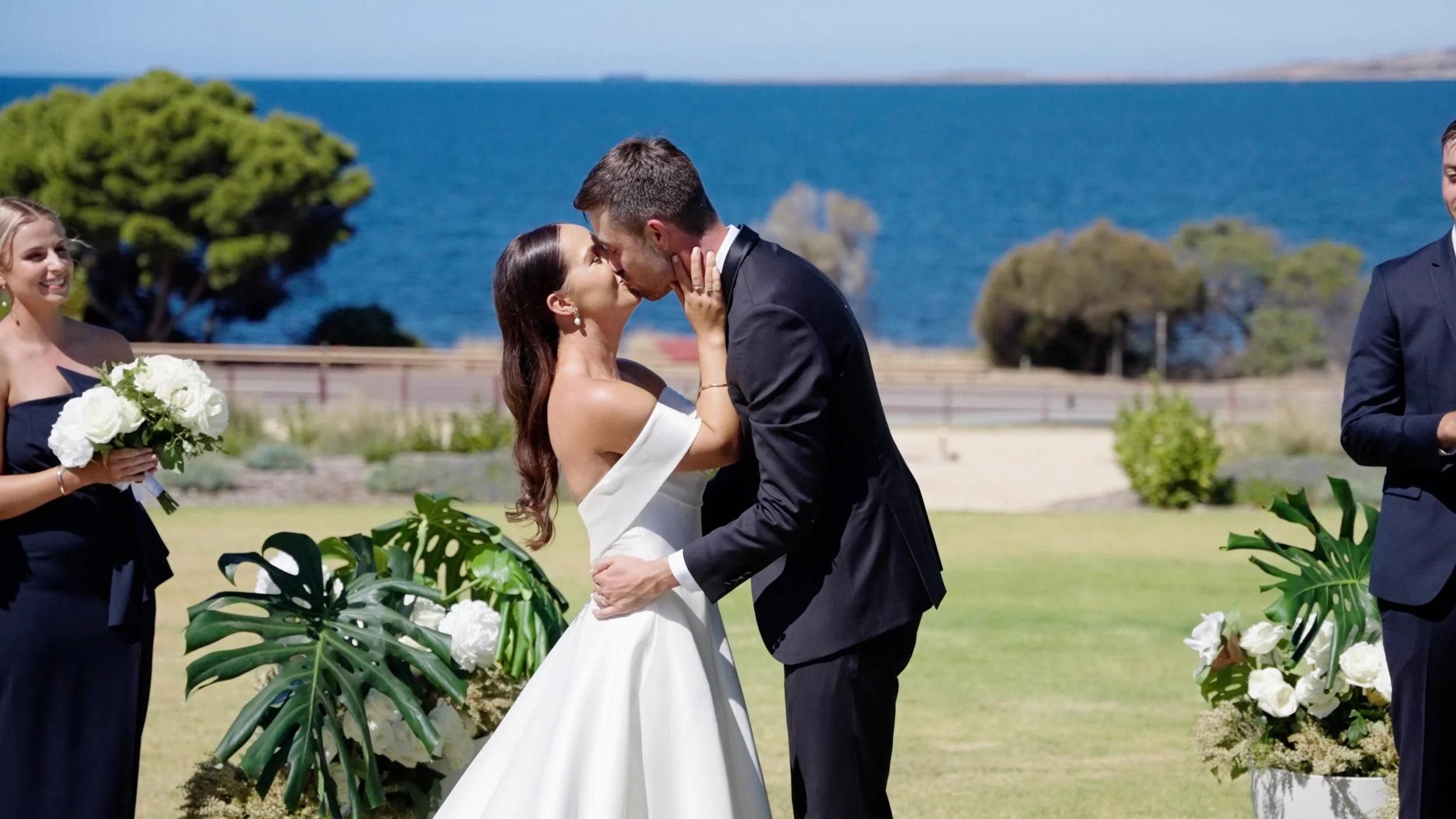 A bride and groom kiss during an outdoor wedding ceremony near the ocean, with bridesmaids and greenery in the background.