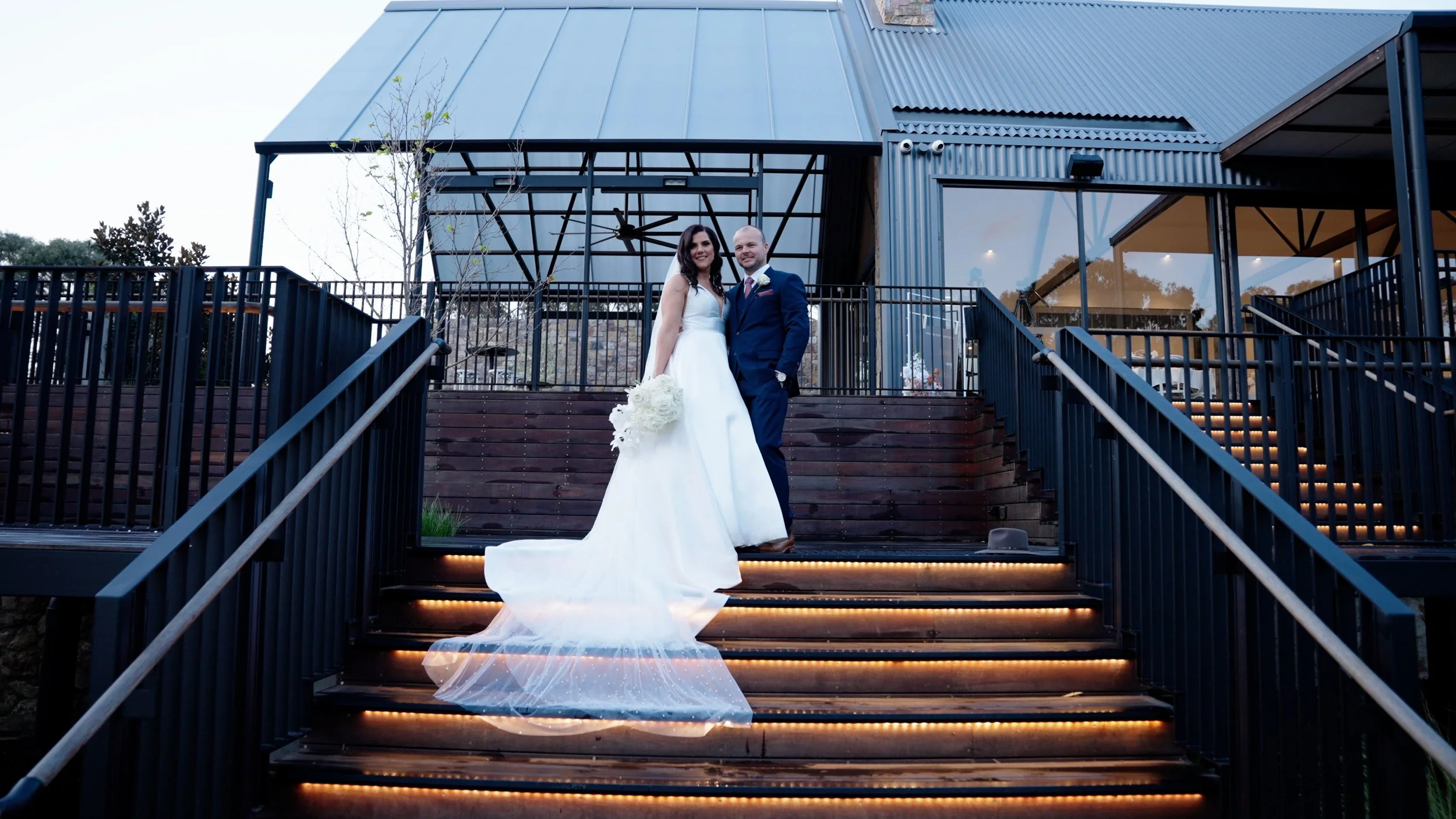 A bride in a white wedding dress holding a bouquet and a groom in a navy suit standing on outdoor wooden stairs with LED lighting, in front of a modern glass and metal building.