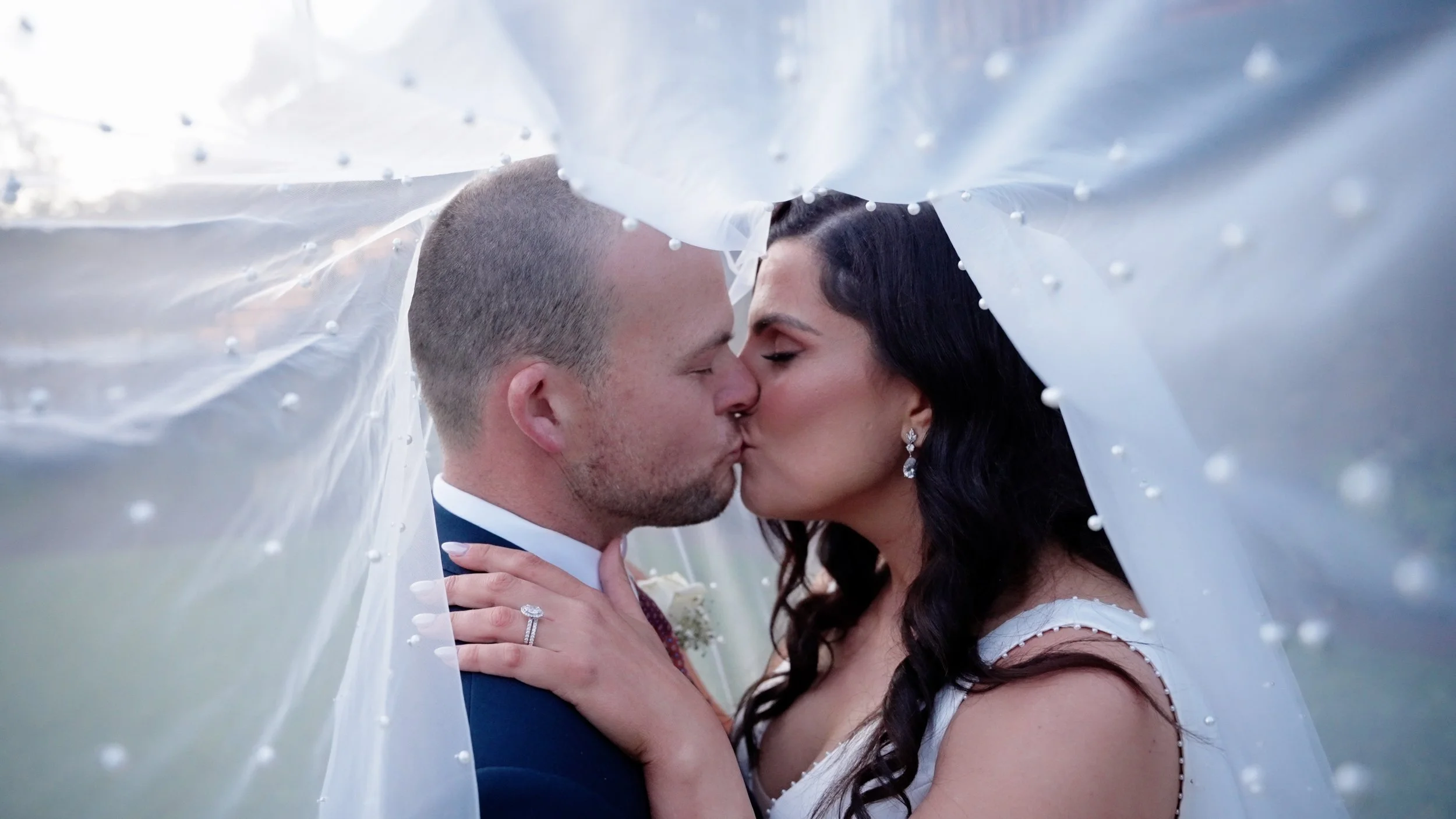 Bride and groom kissing under a wedding veil, outdoors, close-up shot.