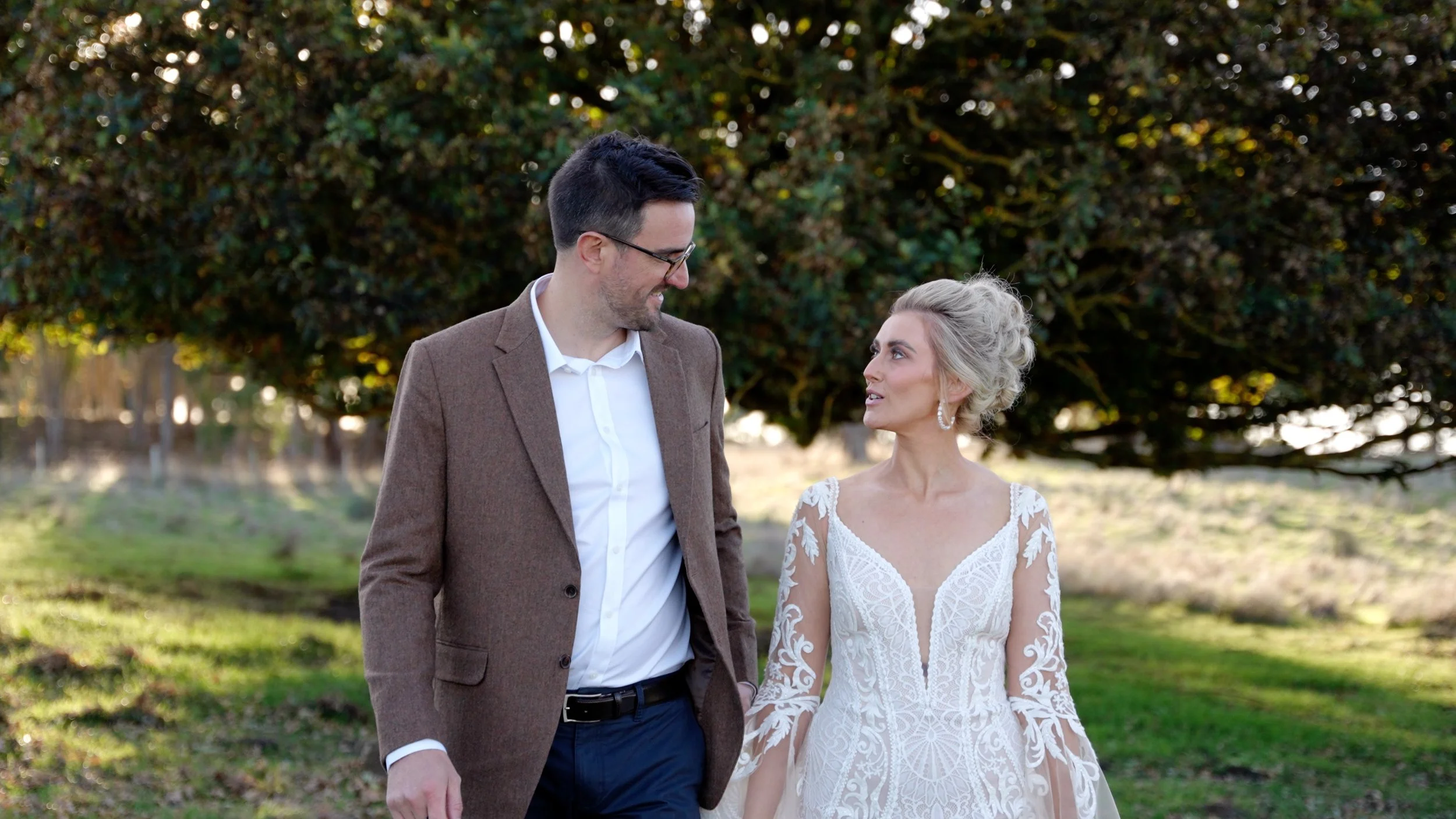 A couple dressed in wedding attire walking outdoors, the man in a brown suit and glasses, the woman in a white lace wedding dress with long sleeves, holding hands and looking at each other with a large tree in the background.