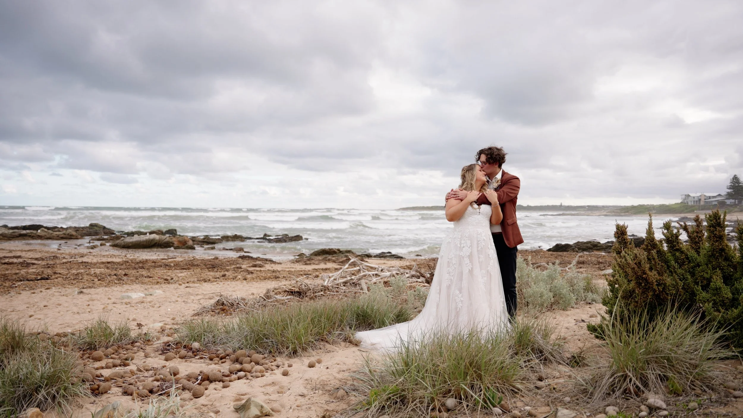 A bride and groom embrace on a beach, with ocean waves and cloudy sky in the background.