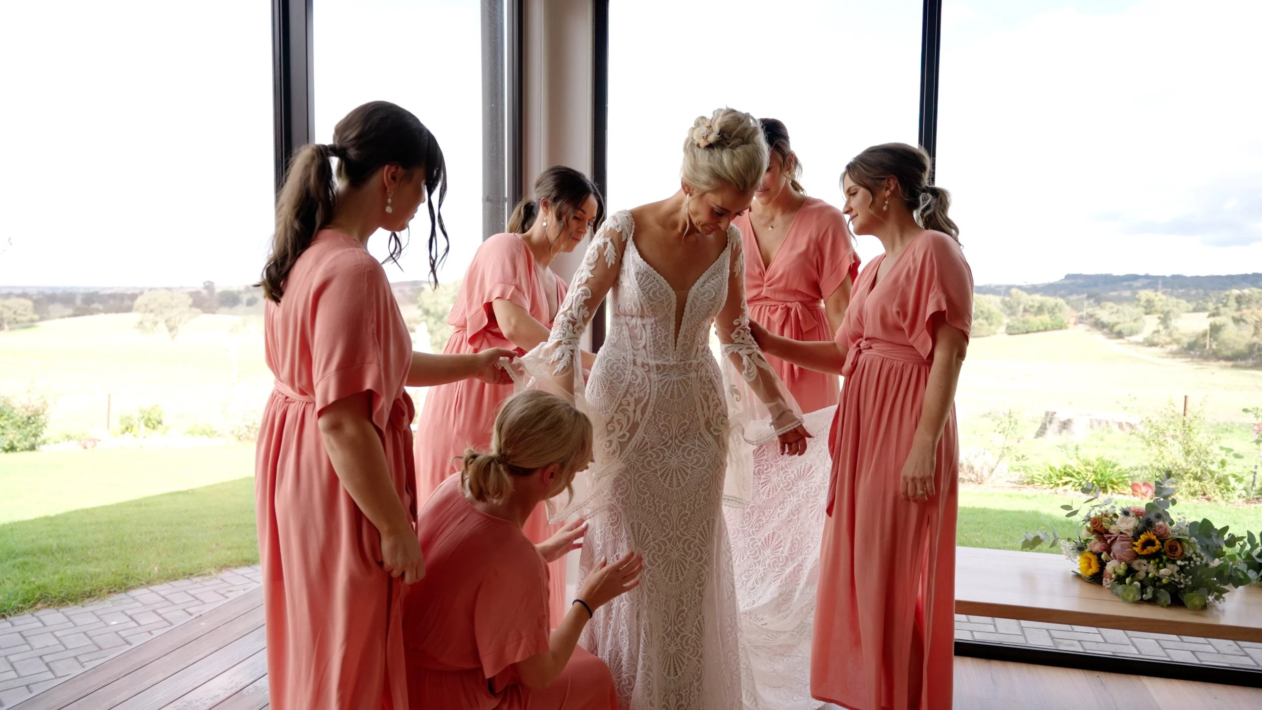 Bride in a white lace wedding dress surrounded by bridesmaids in pink dresses, helping her with her dress indoors with large windows and a scenic outdoor view.