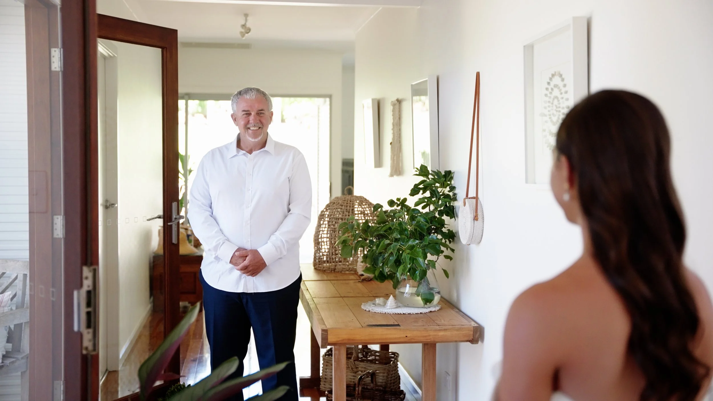 A man in a white shirt smiling and standing in a bright doorway, while a woman with long hair is seated inside, facing him.