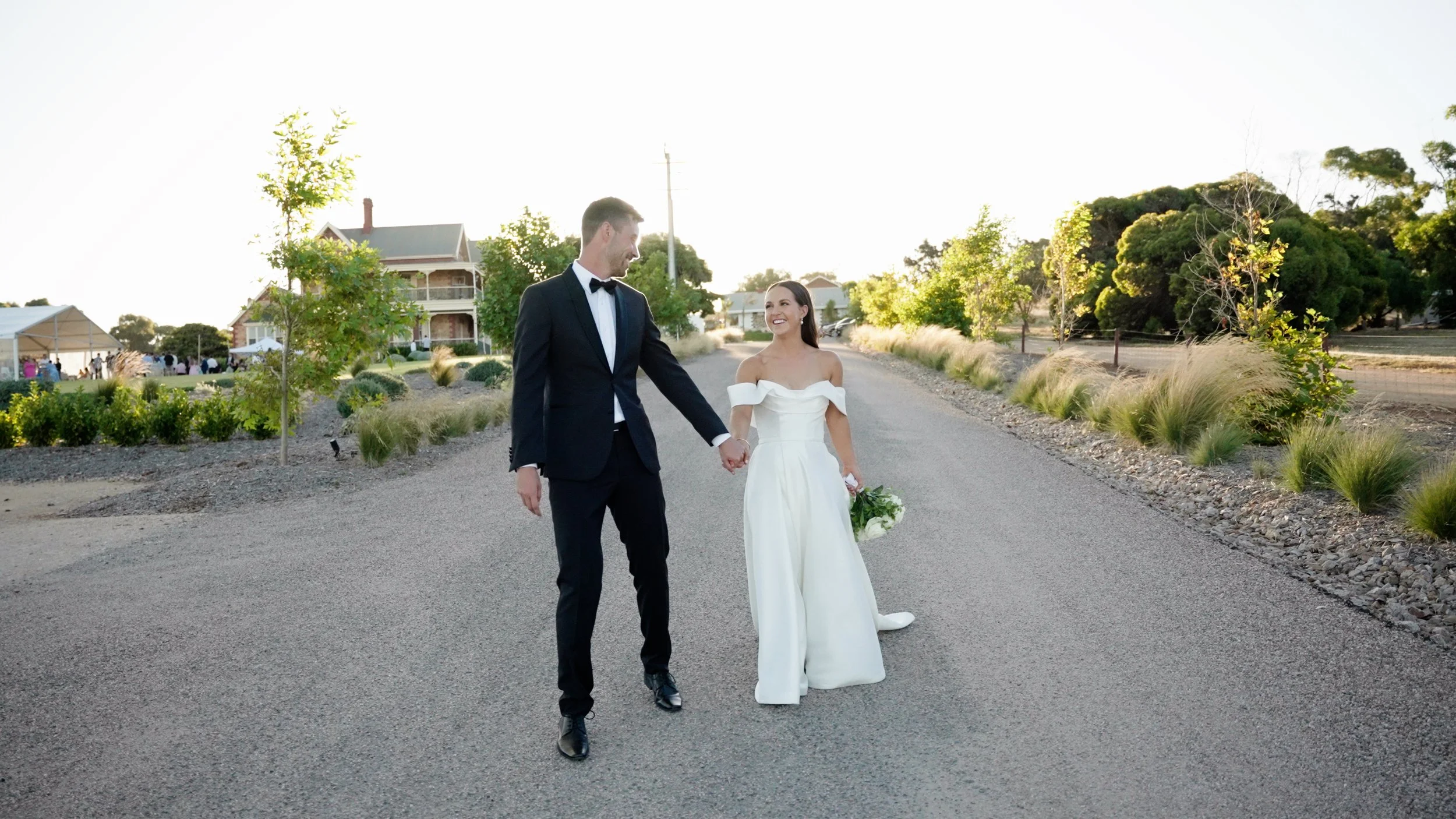 A bride and groom in wedding attire walking hand in hand on a paved road, surrounded by landscaped greenery and trees, with a house and a small crowd in the background.