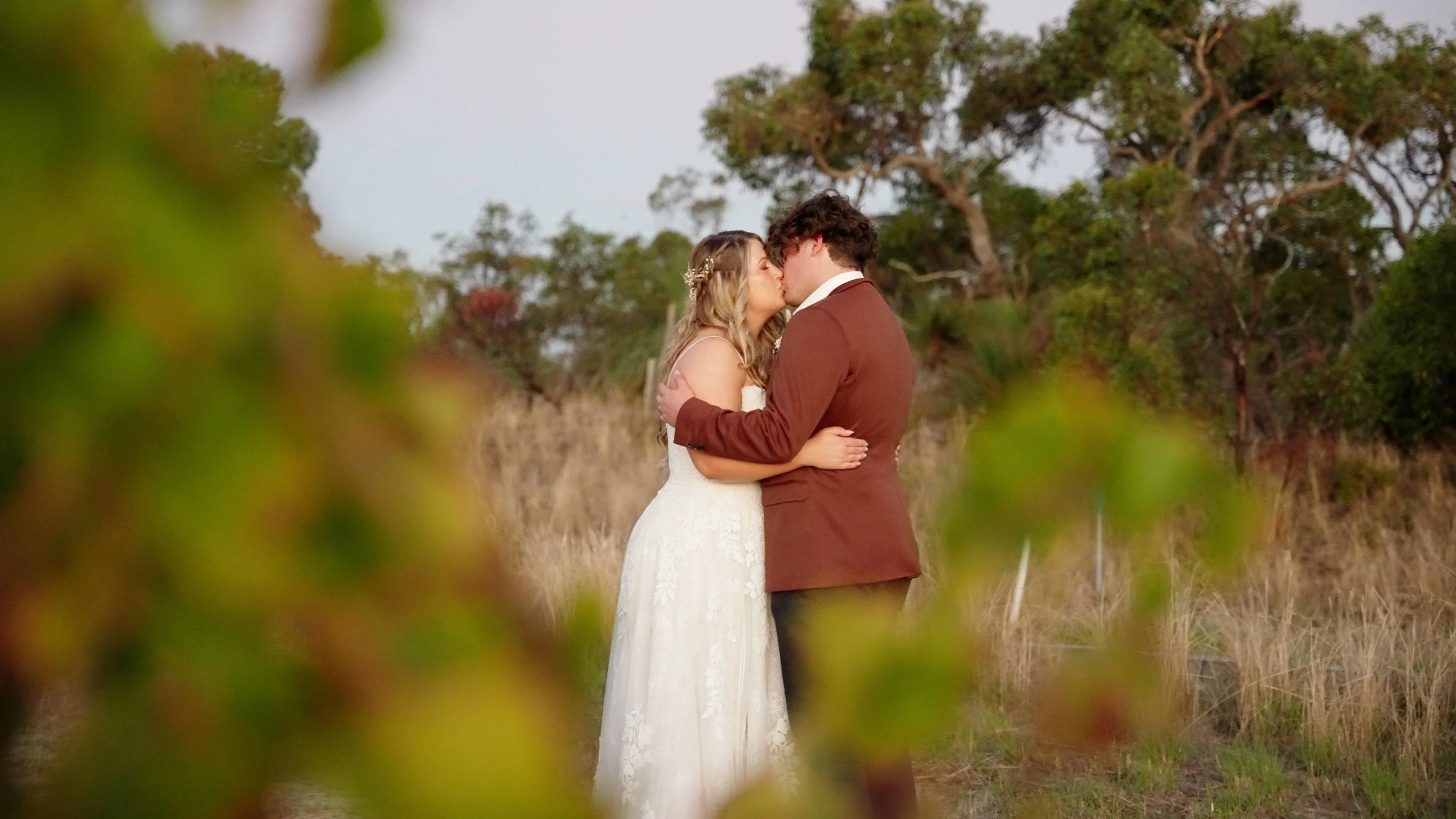 A bride and groom kissing outdoors, framed by blurred green leaves, in a grassy field with trees in the background.