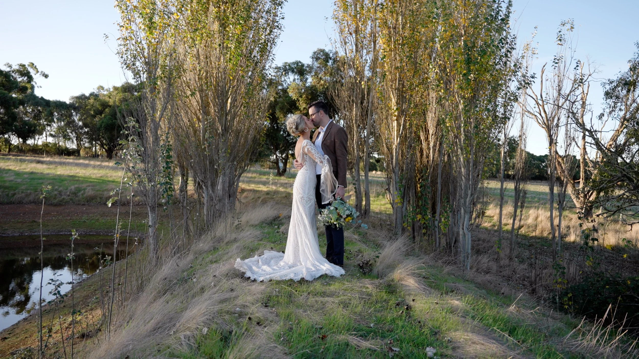 Couple in wedding attire standing on a grassy path surrounded by trees, about to kiss, outdoors in natural light