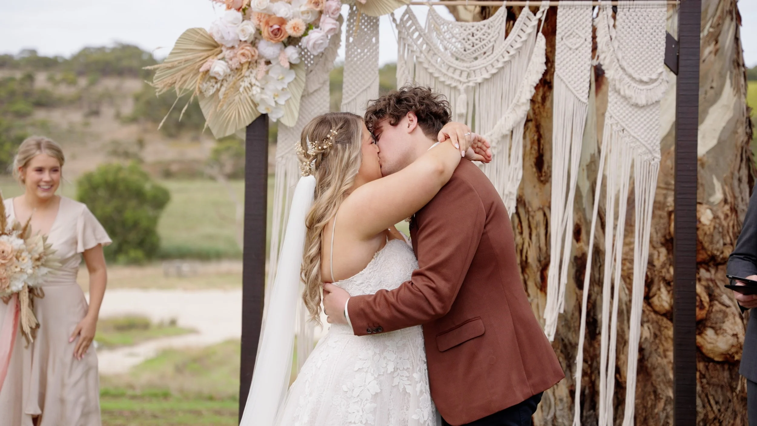 A newlywed couple sharing a kiss during their outdoor wedding ceremony under a decorated arch, with a bridesmaid smiling in the background.