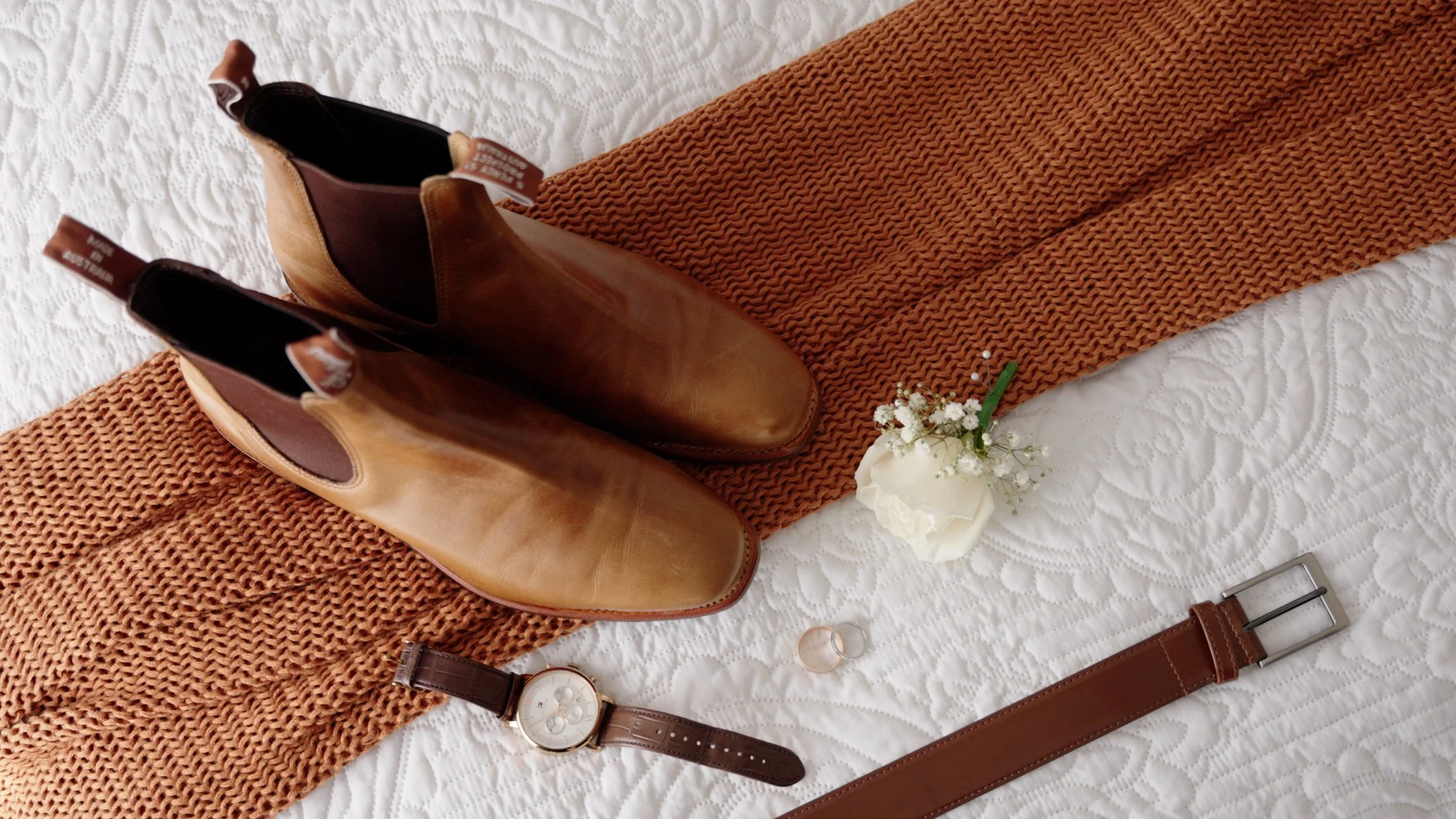 Pair of brown leather ankle boots, a wristwatch, a wedding band, a small bouquet of white flowers, and a brown leather belt laid on a bed with a white quilt and a brown knitted scarf.