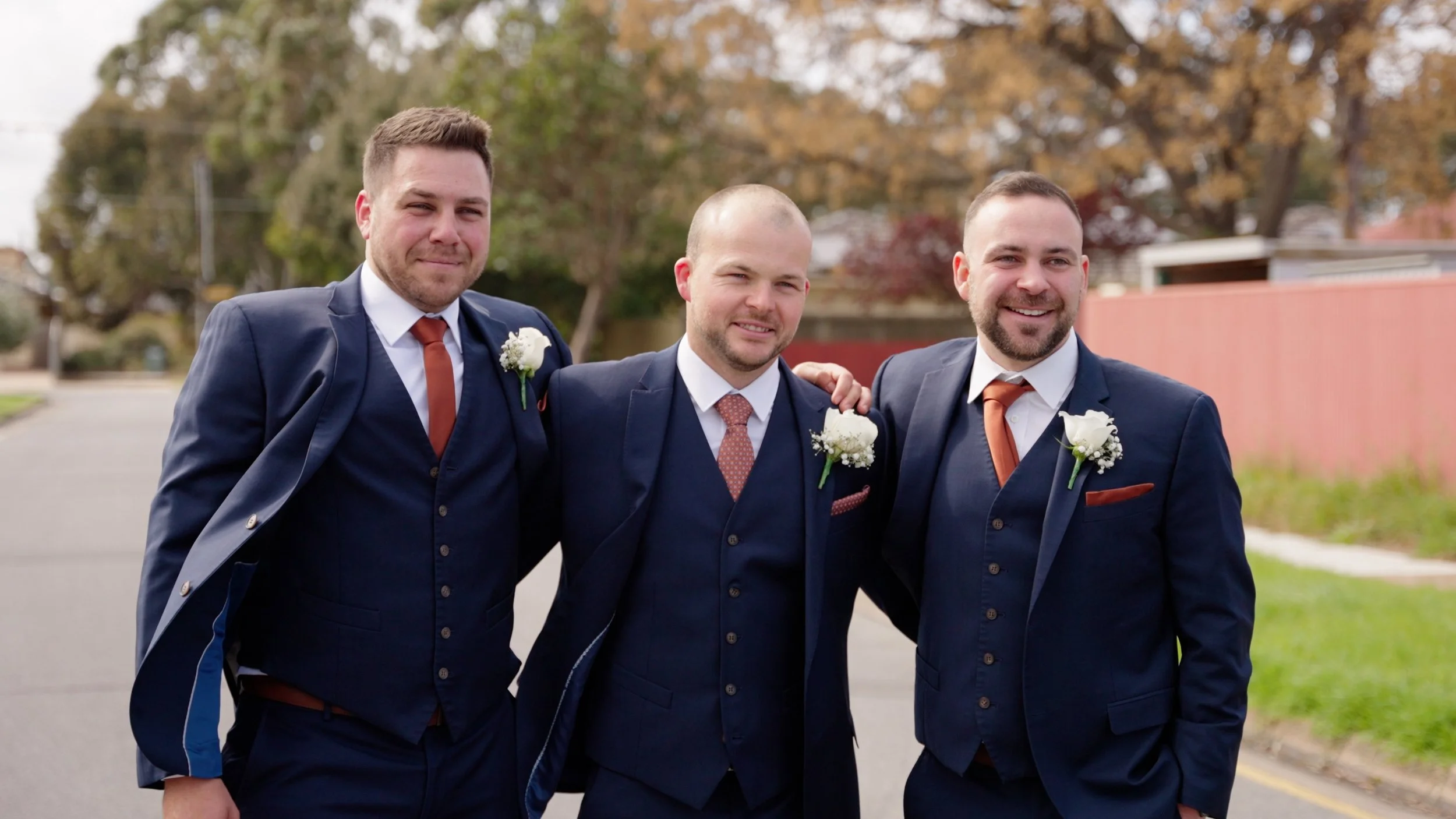 Three men dressed in navy suits and ties standing outside on a street, smiling, with one man in the center and the other two on either side, all with flower boutonnieres. They appear to be at a wedding.