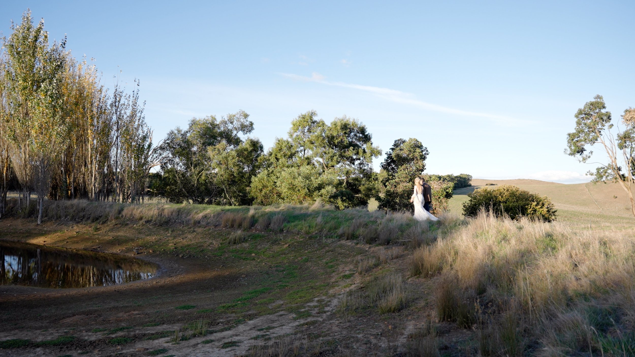 A bride and groom standing together in a rural outdoor setting with trees and grassy hills, reflecting in a small pond nearby.