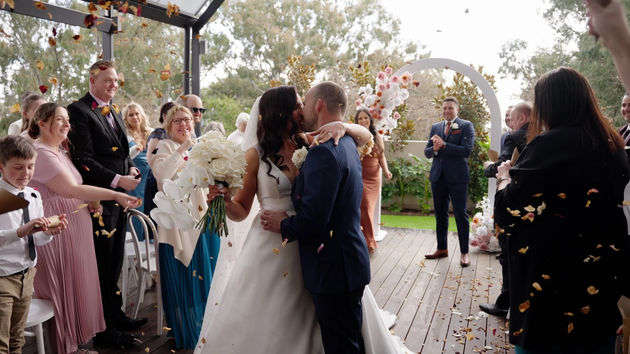 A newly married couple shares a kiss under a floral arch as friends and family celebrate with confetti at an outdoor wedding ceremony.
