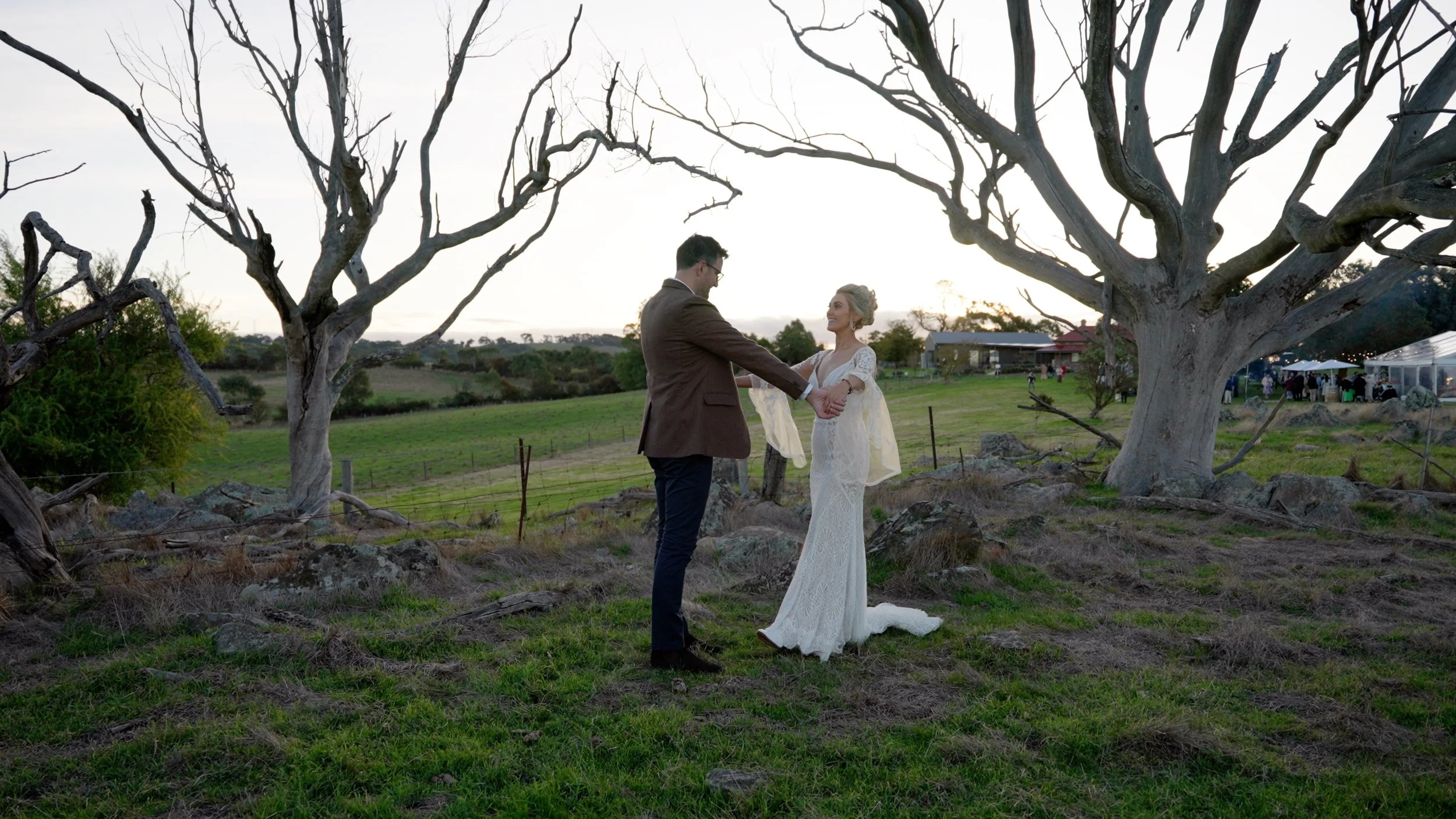 A couple holding hands and smiling at each other outdoors in front of two large leafless trees during sunset, with a grassy field and a distant building in the background.