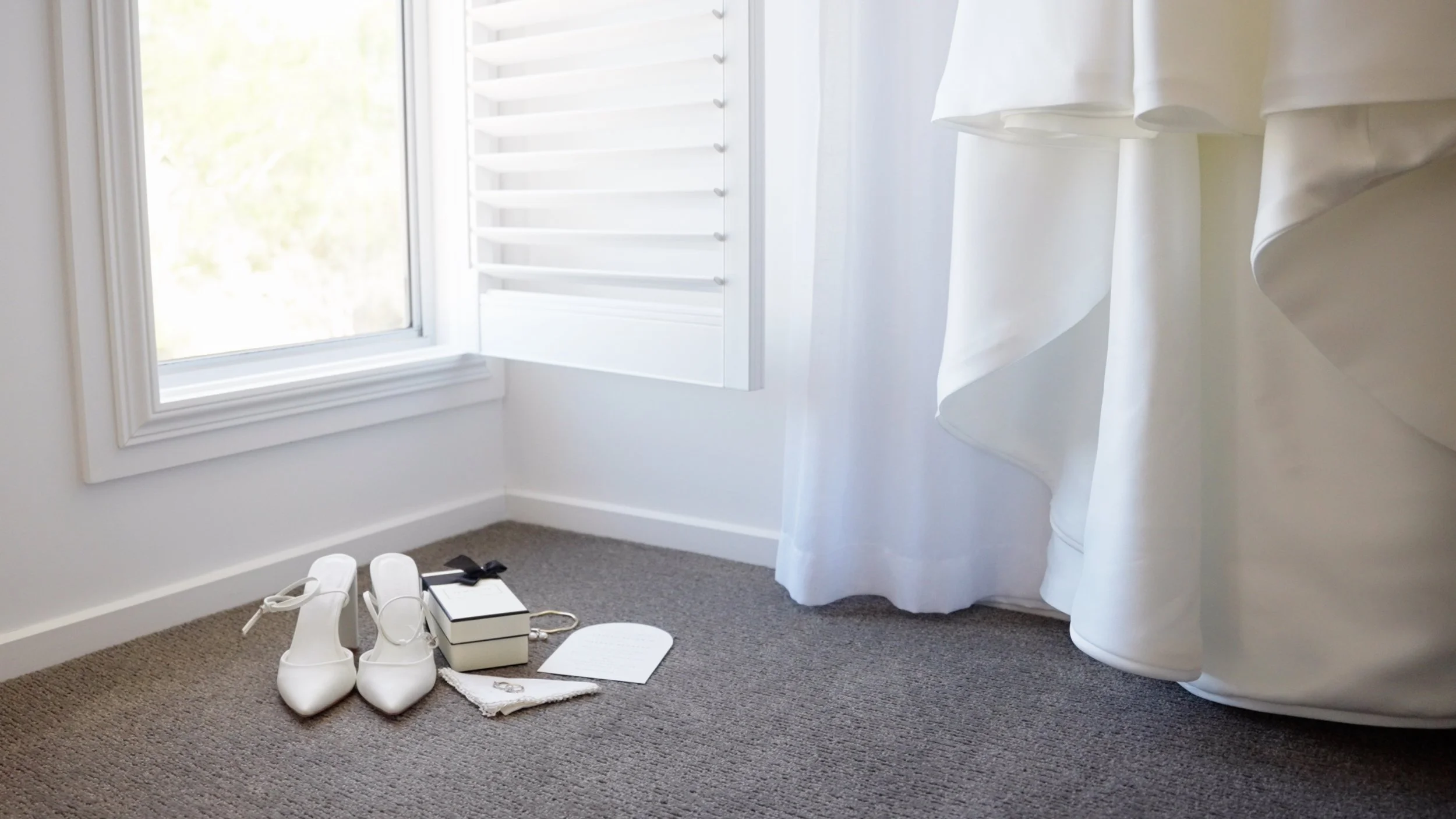 White wedding shoes, a ring, a wedding dress, a small box, and a letter on a beige carpeted floor near a window.