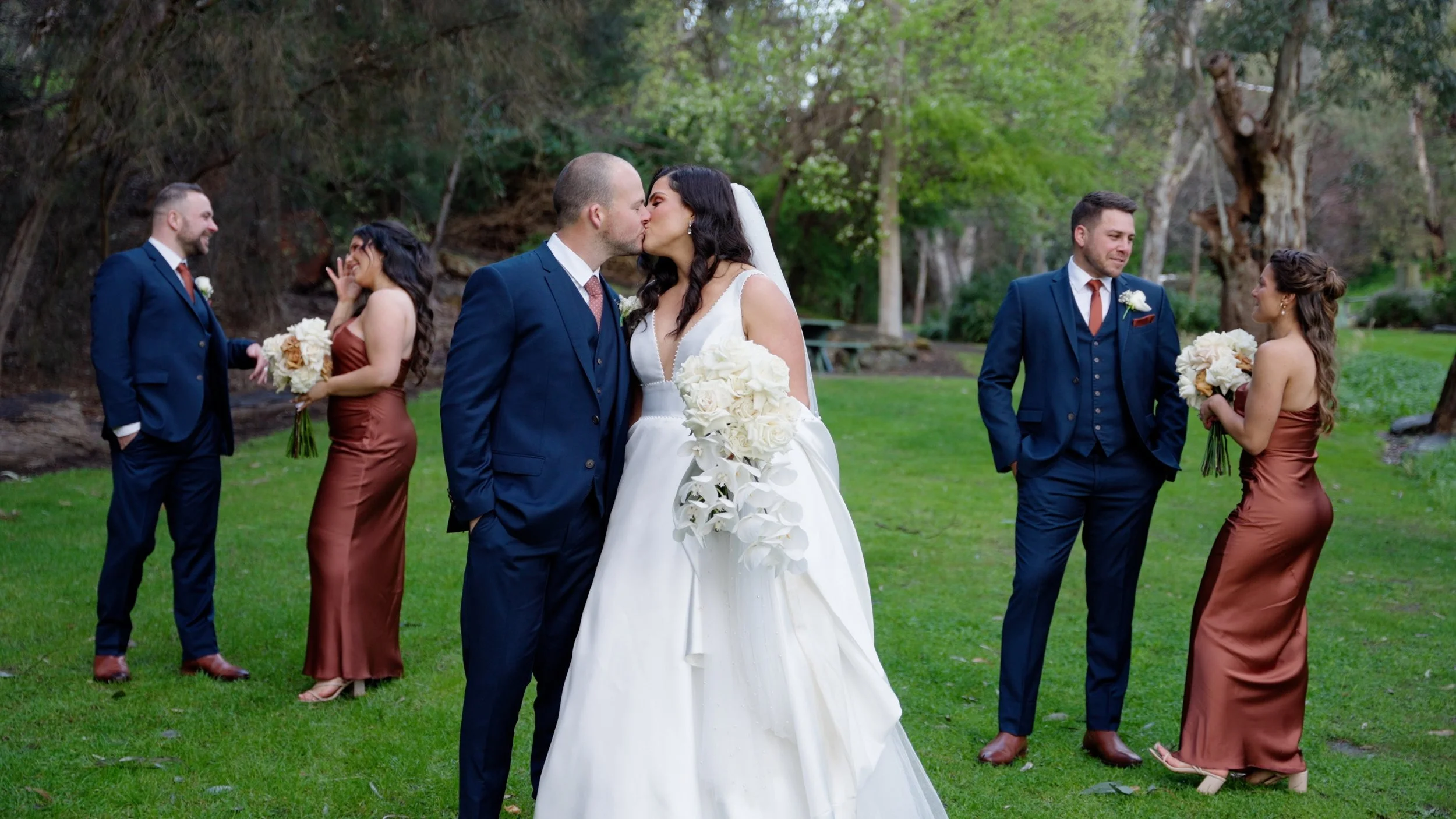 Bride and groom kissing with two bridesmaids and two groomsmen standing on a grassy outdoor area with trees in the background.