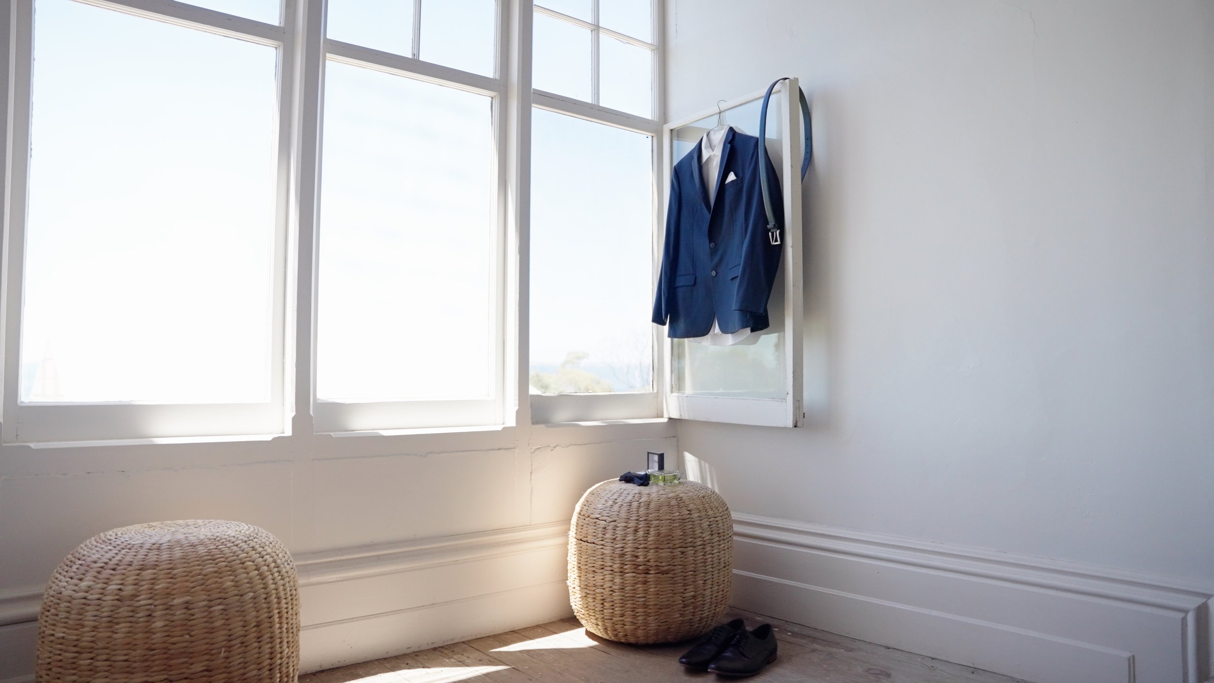 A bright room with large windows, a blue suit jacket hanging on a wall hook, a pair of black shoes, two woven ottomans, and sunlight streaming in.