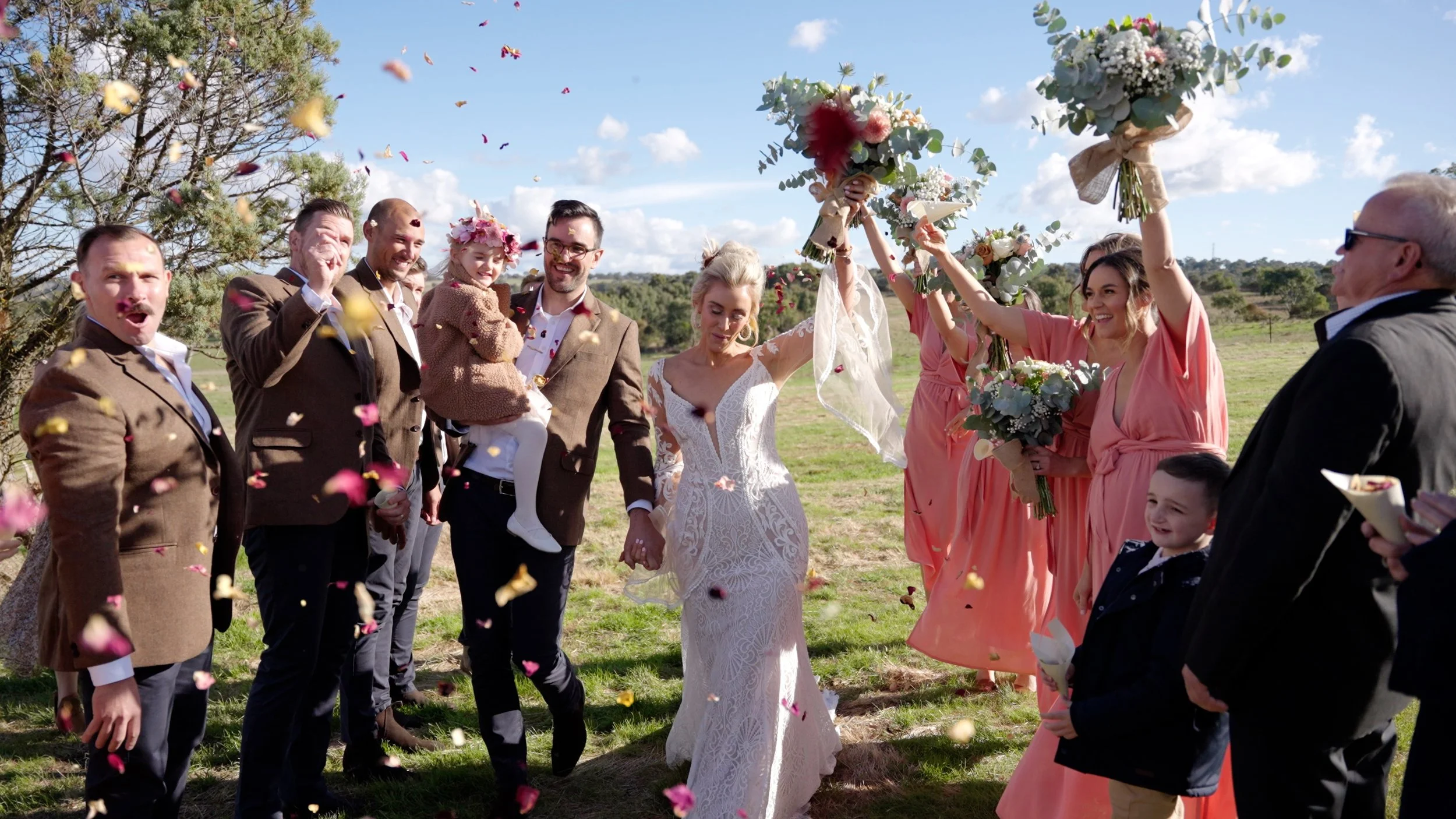 A wedding celebration outdoors with the bride and groom holding hands, surrounded by friends and family throwing flower petals in the air on a sunny day.